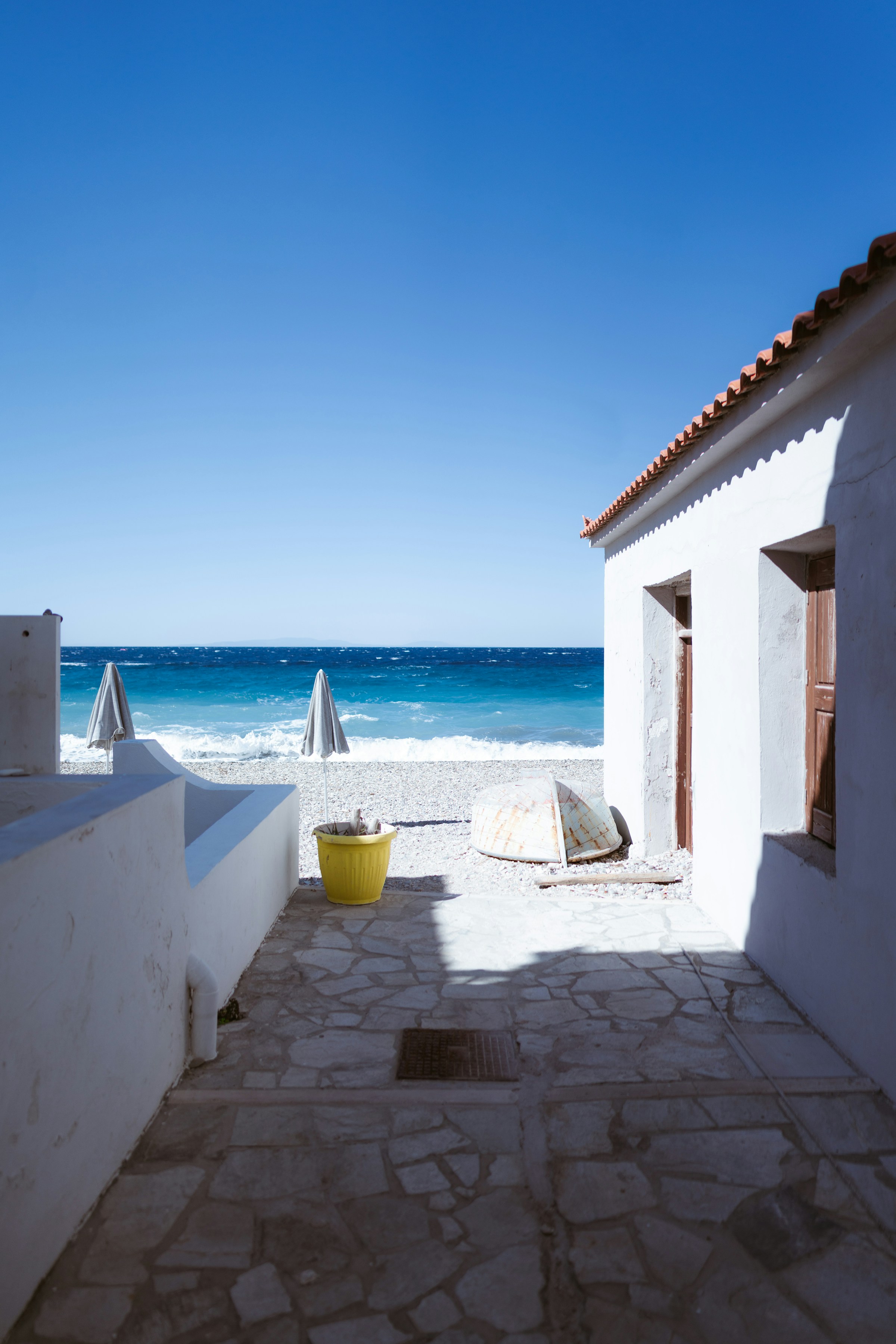 White stone pathway leading to a pebbled beach with blue ocean waves and umbrellas, set against a clear sky. A white building with a red-tiled roof.