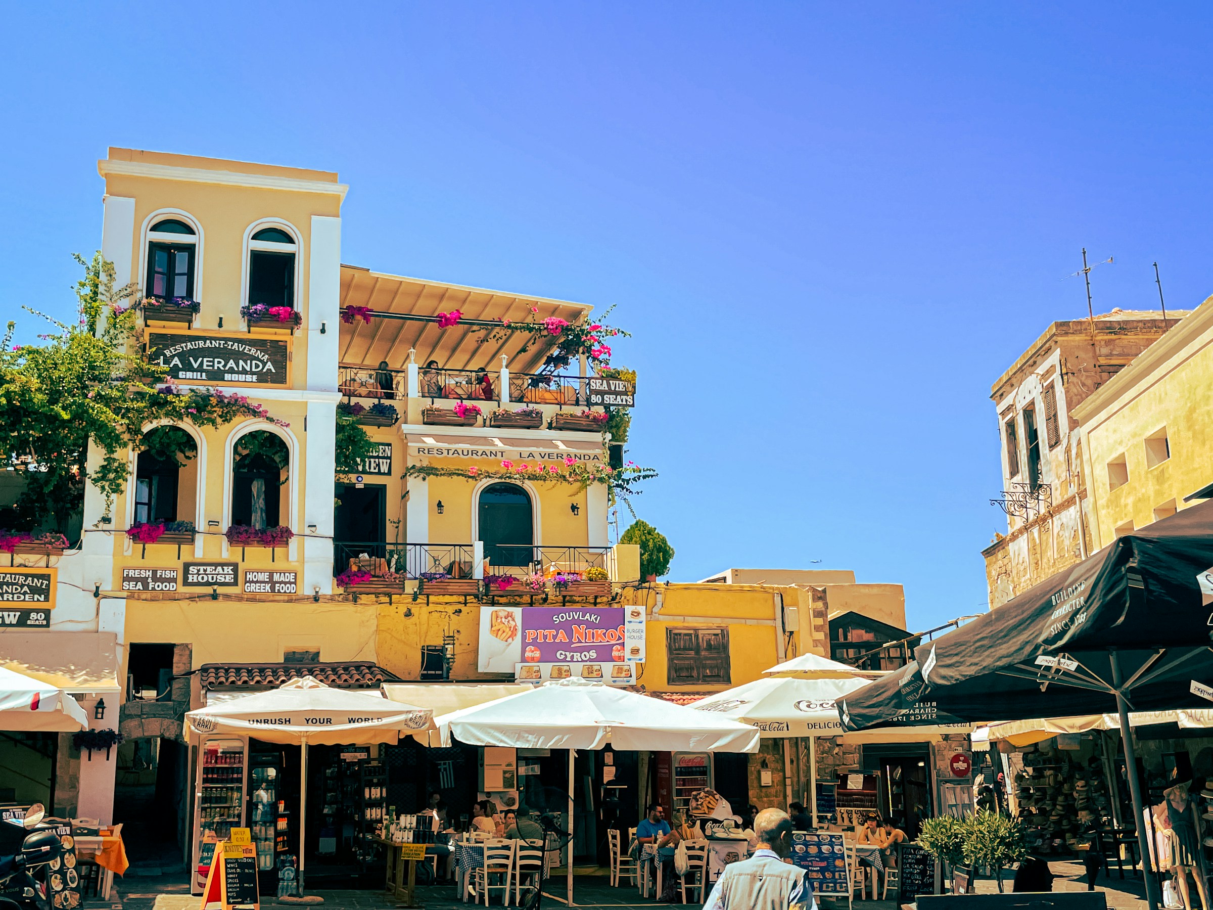 Historic Mediterranean building with colorful flower boxes and outdoor cafes under sunny blue skies.