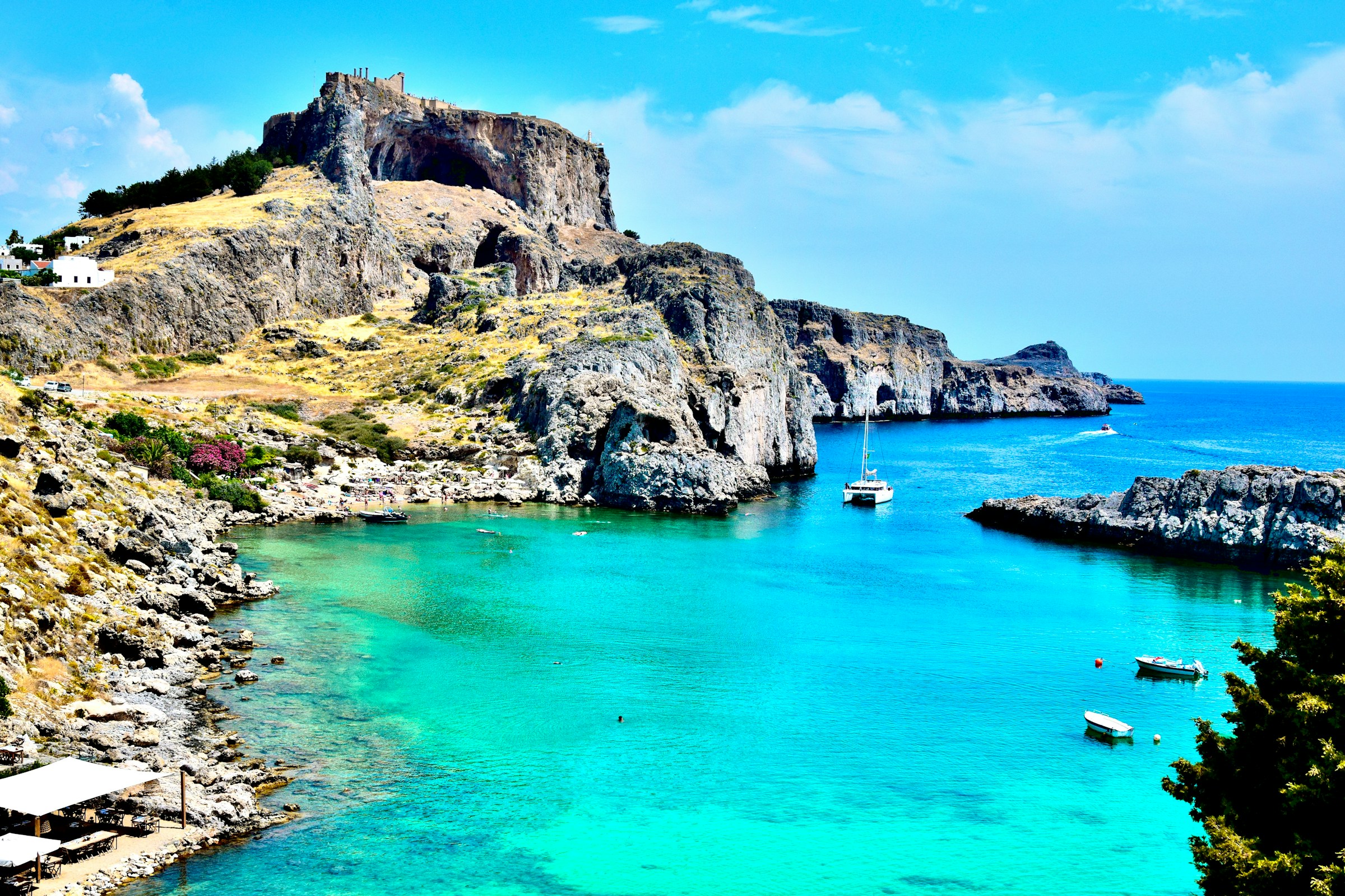 Scenic view of turquoise bay surrounded by rocky cliffs with sailboats anchored in crystal-clear water under a clear blue sky.