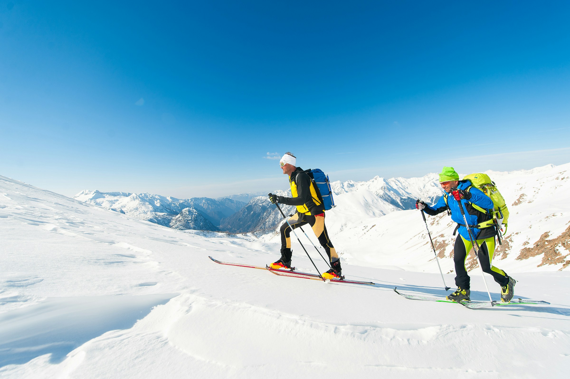 To mennesker på topptur med skiutstyr, klatrer opp en snødekt fjellside under en klar blå himmel. Majestetiske fjell i bakgrunnen
