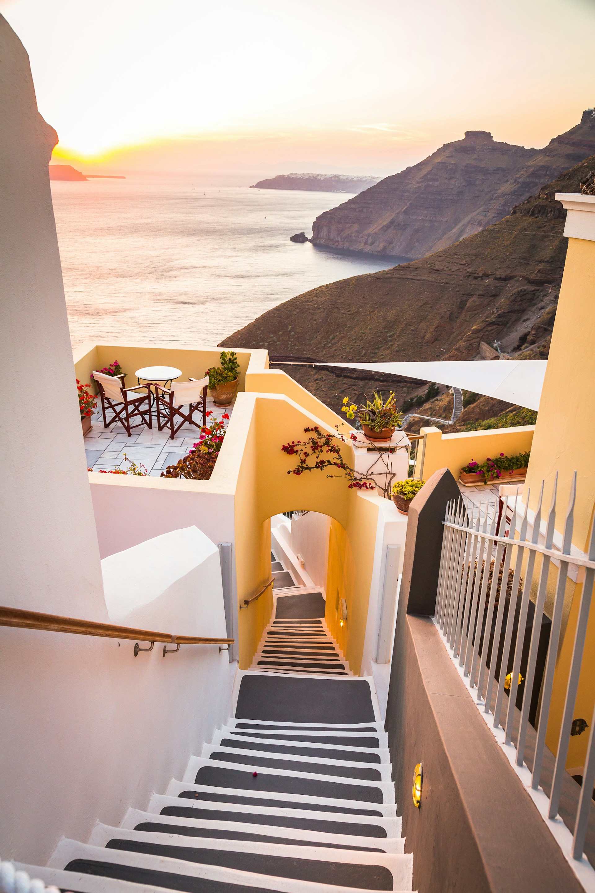 White and yellow stairway leading to a terrace with ocean view in Santorini at sunset, featuring potted plants and distant cliffs.