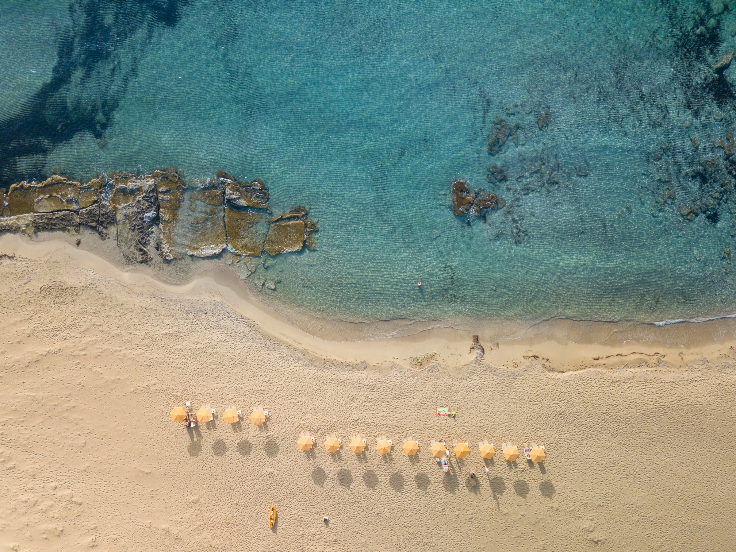 Aerial view of a sandy beach with clear turquoise water, featuring yellow beach umbrellas and lounge chairs lined up near the shoreline. Rocky formations are visible in the water.