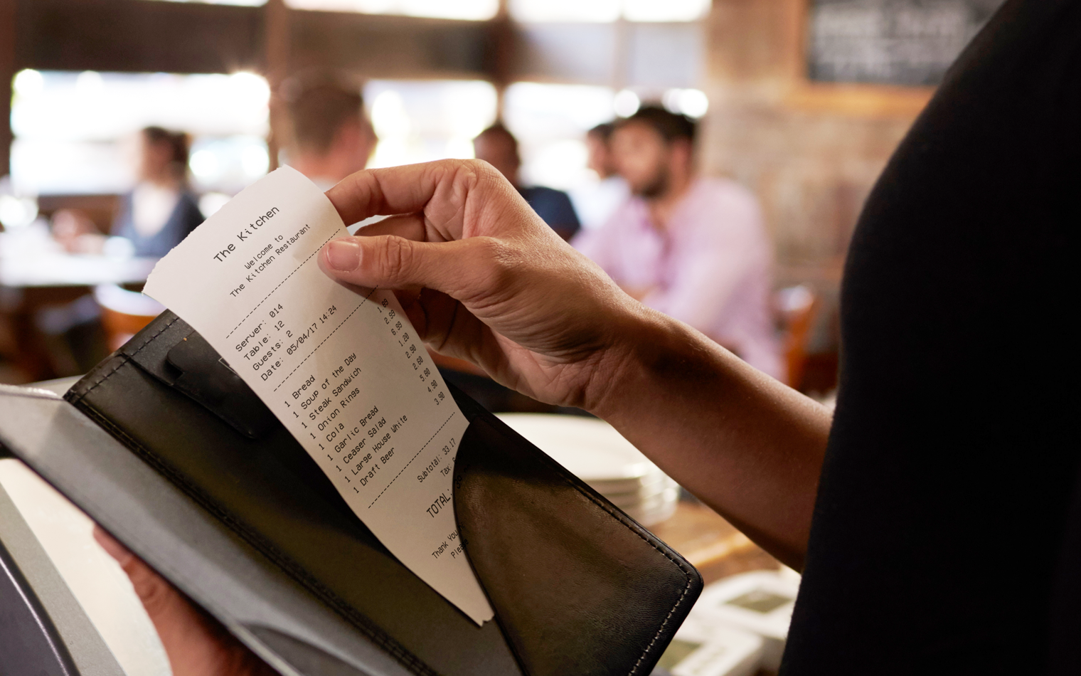 Personen som holder en restaurantregning i en skinnholdere mens de sitter ved et bord på en restaurant. Flere personer kan sees i bakgrunnen, samt et uklart restaurantinteriør.