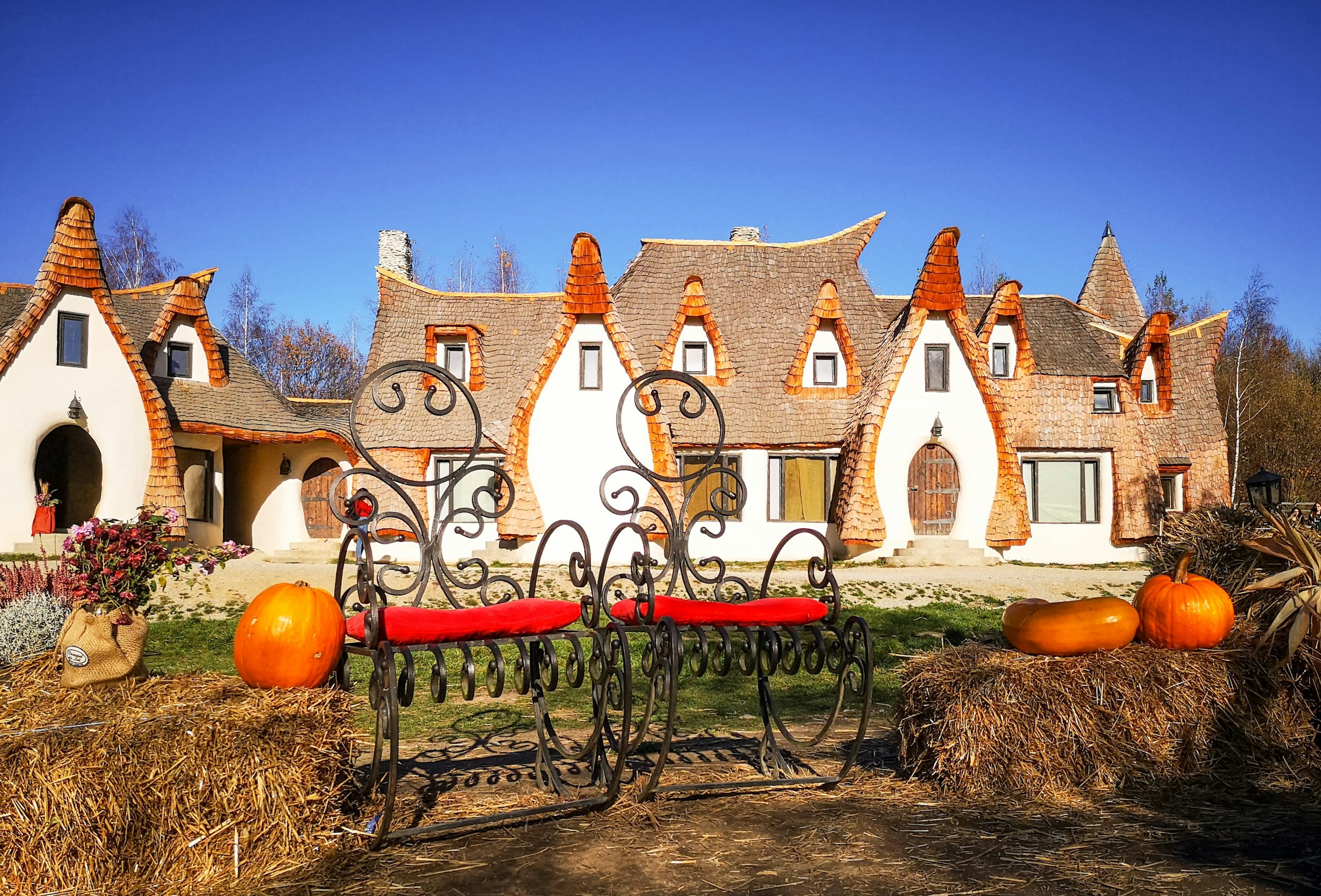 Fabelaktig arkitektur med spirtak av et uvanlig hus, smijernsbenker og høstlige gresskar foran bygningen under en klar blå himmel.