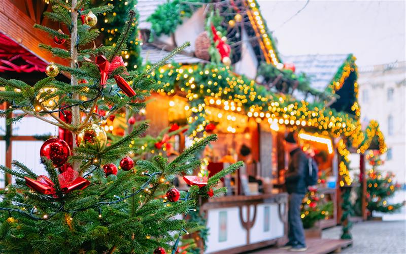 Christmas tree decorated with red baubles and lights in the foreground at an atmospheric Christmas market with decorated market stalls in the background.