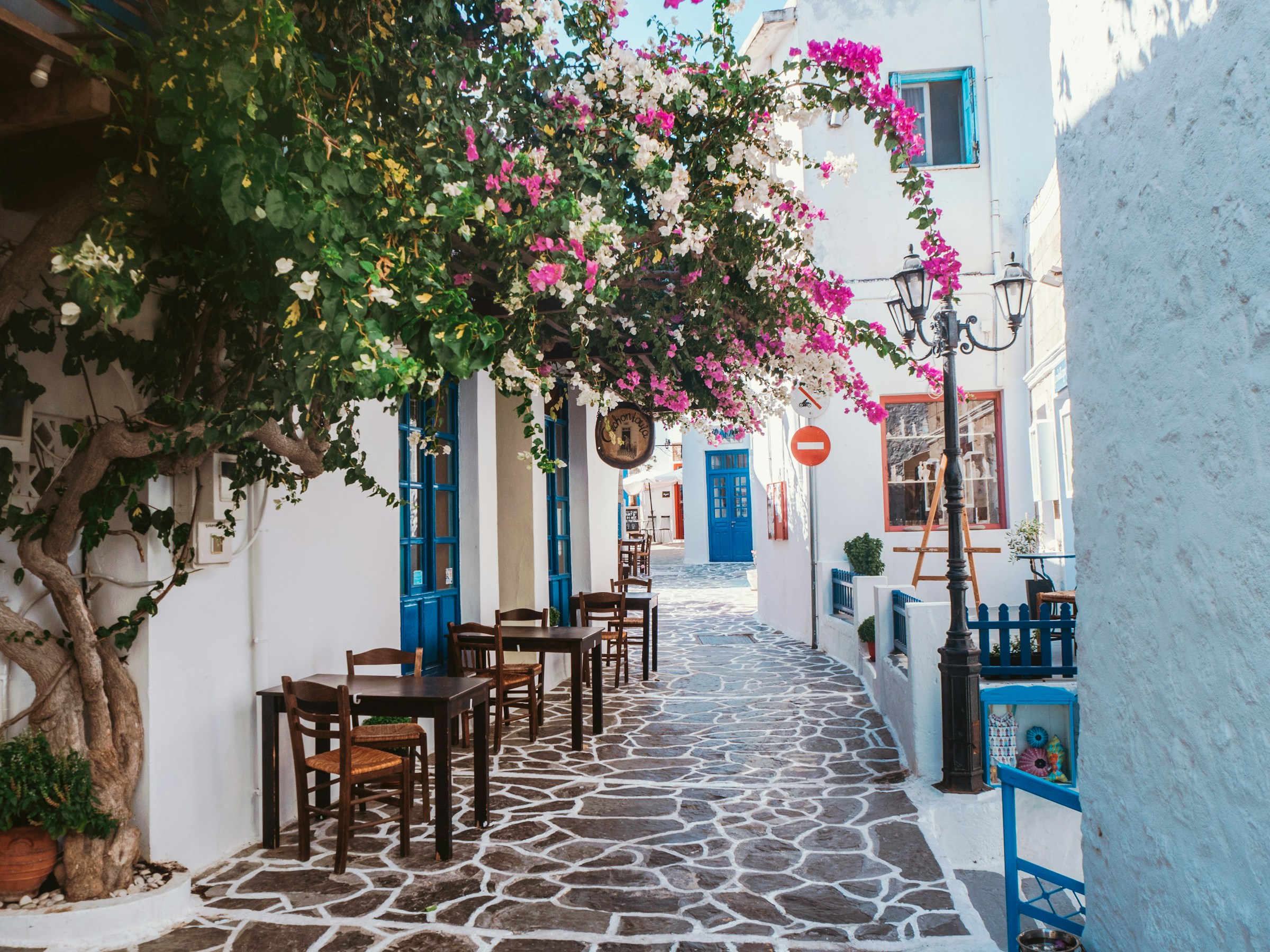 Charming narrow street in a Greek village with blue and white buildings, vibrant bougainvillea, and outdoor seating