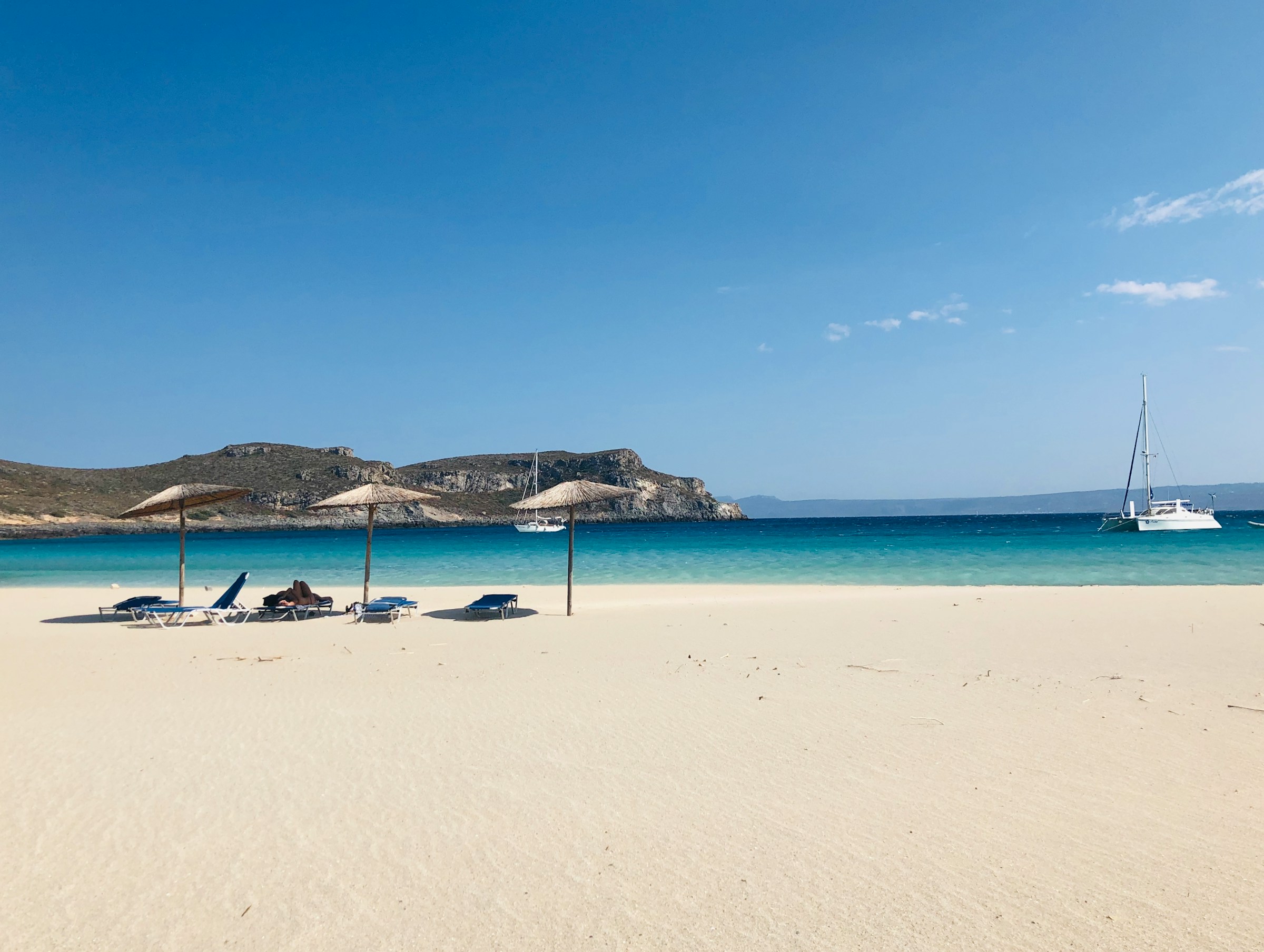 Sandy beach with sun loungers and straw umbrellas facing turquoise sea, with a sailboat in the distance against a clear blue sky