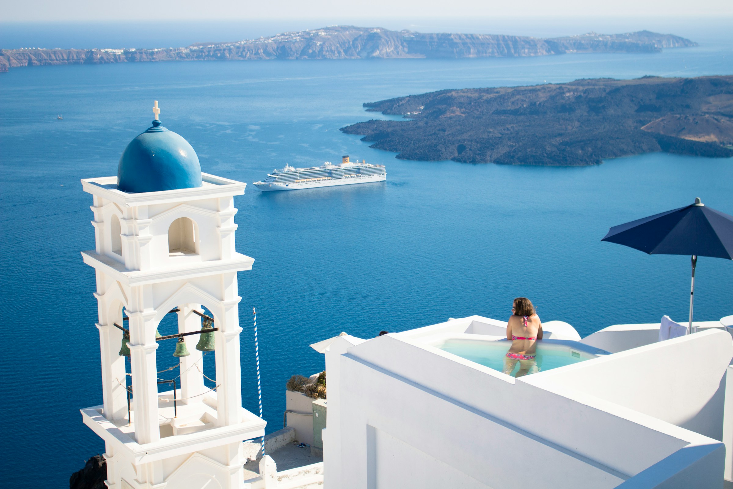 Woman relaxing in an infinity pool overlooking the sea with a cruise ship and blue-domed church in Santorini, Greece