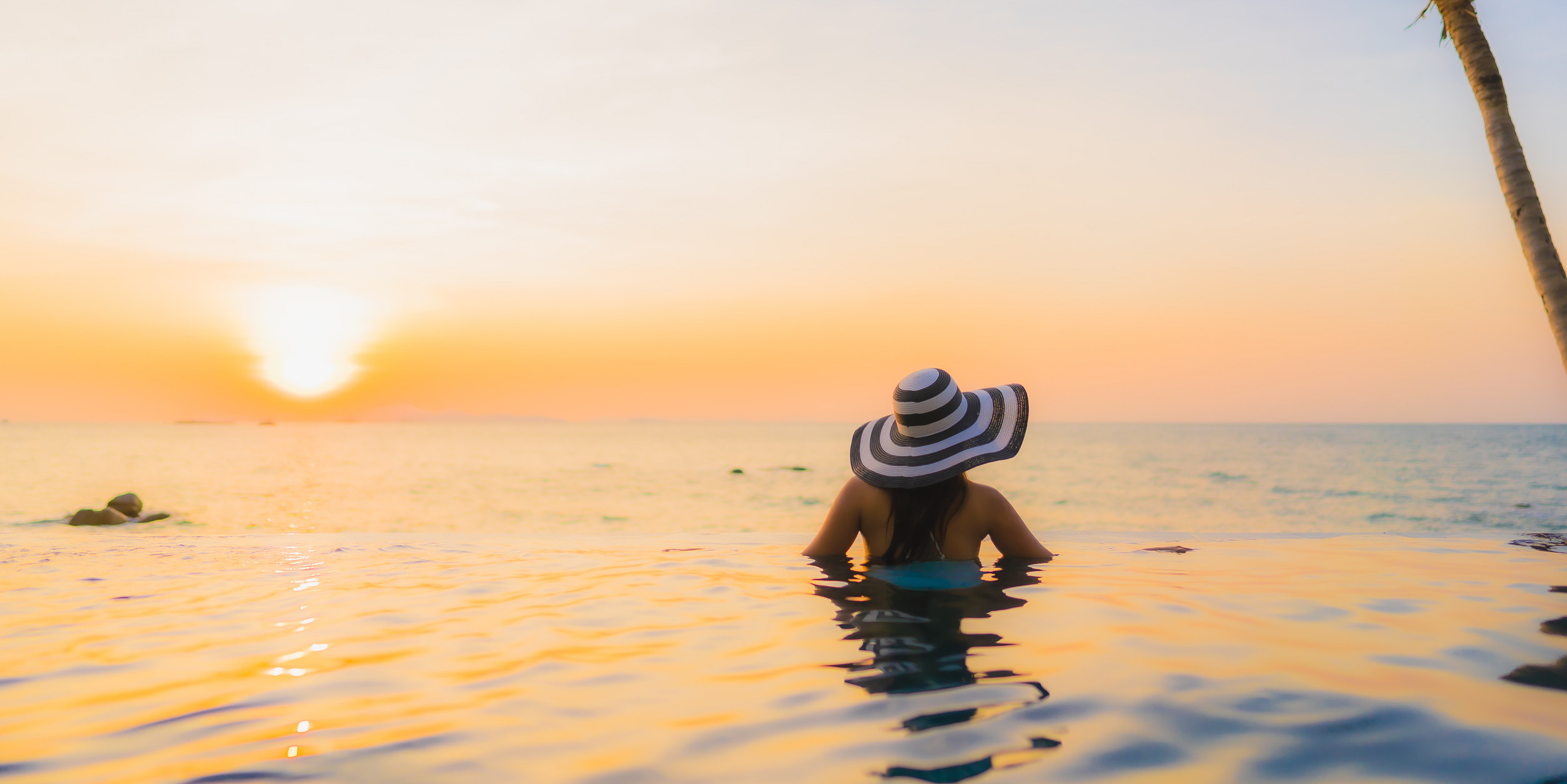 Woman in a wide-brimmed hat relaxing in an infinity pool at sunset, overlooking the ocean.