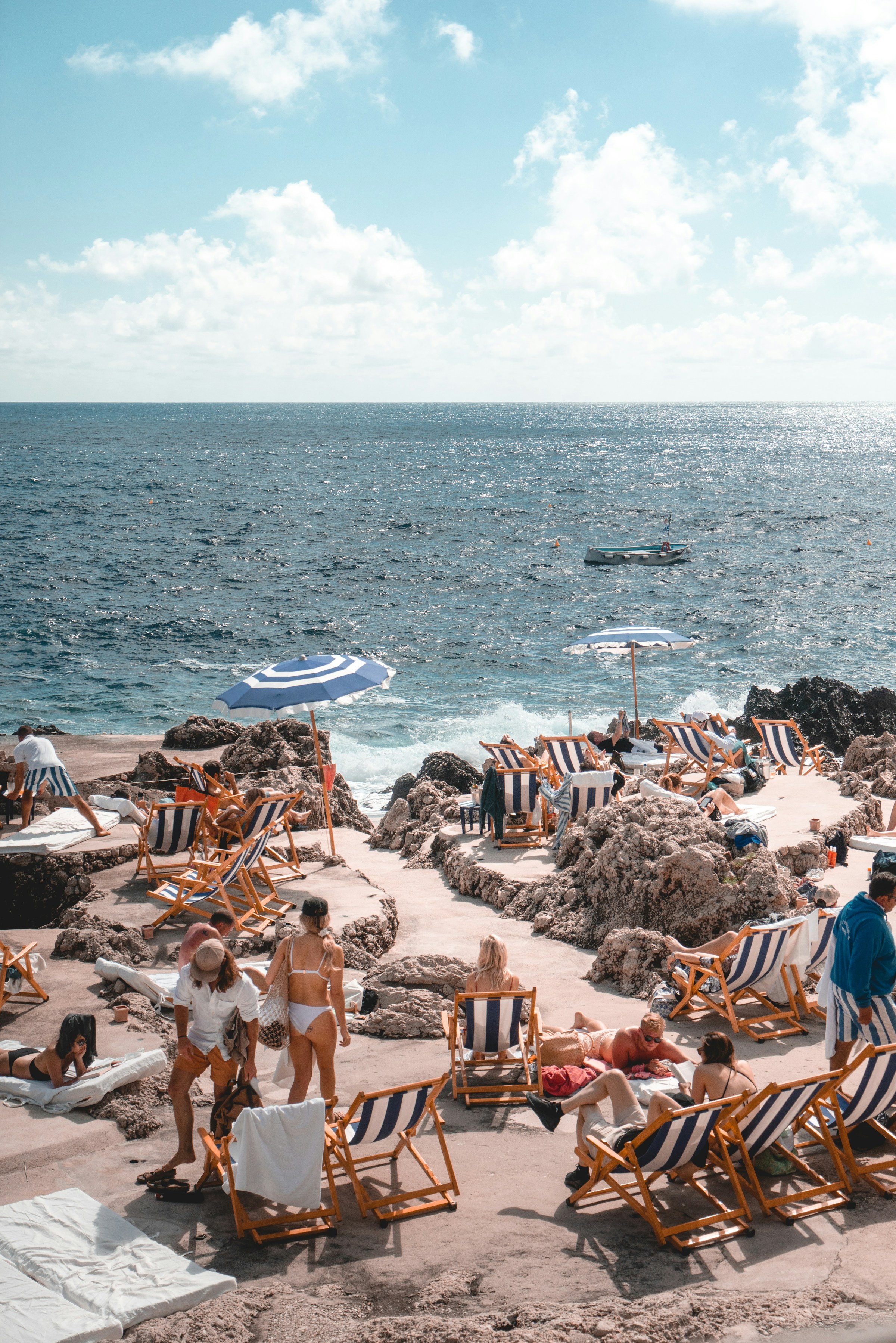 Solsenger og parasoller på den steinete kysten med turkis hav og badende som nyter strandferie ved Middelhavet. Avslappende strandmiljø i Sør-Europa.