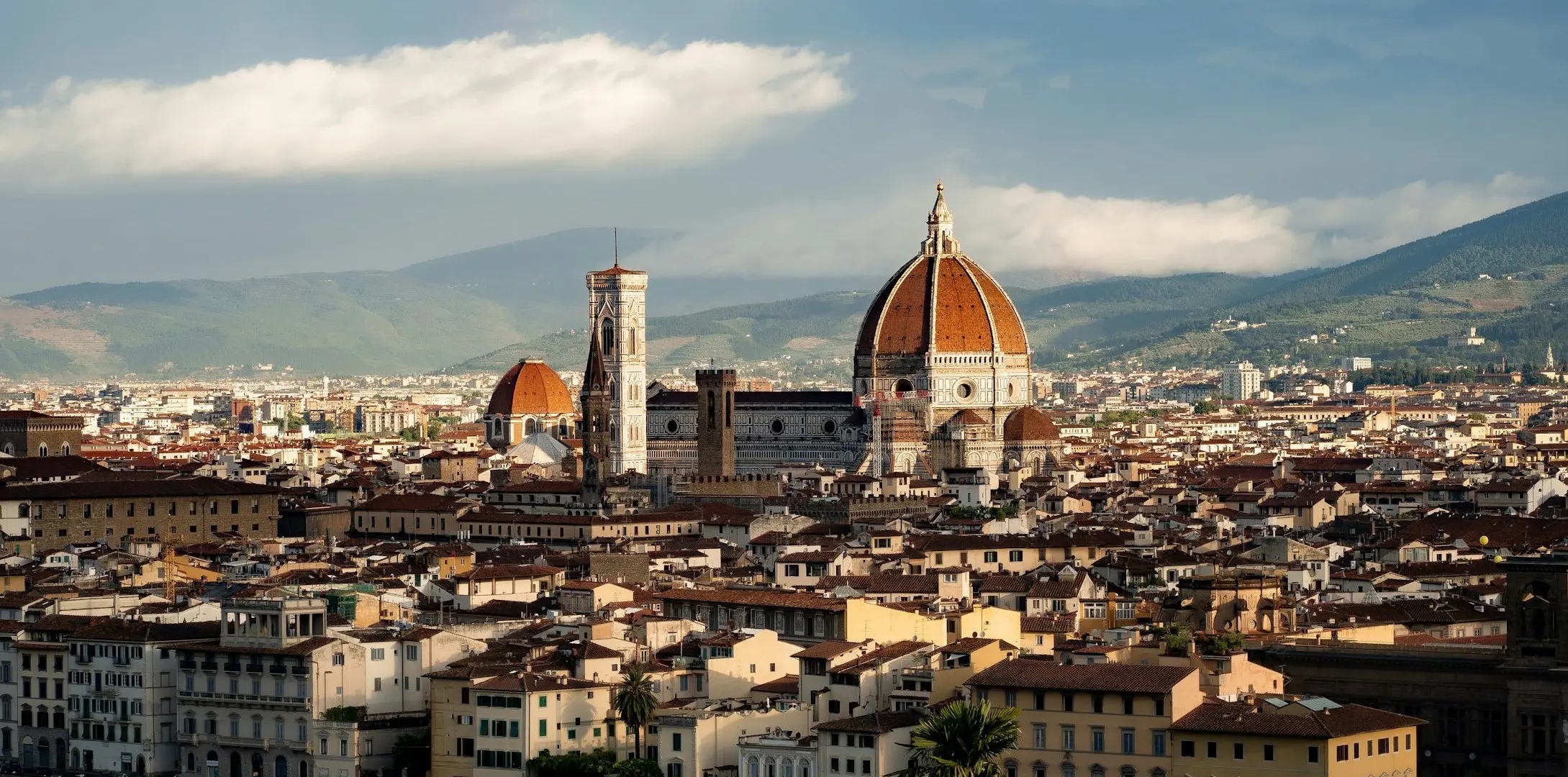 Panoramic view of Florence, Italy, featuring the iconic Florence Cathedral with its red-tiled dome, surrounded by historic city buildings and distant hills under a partly cloudy sky.