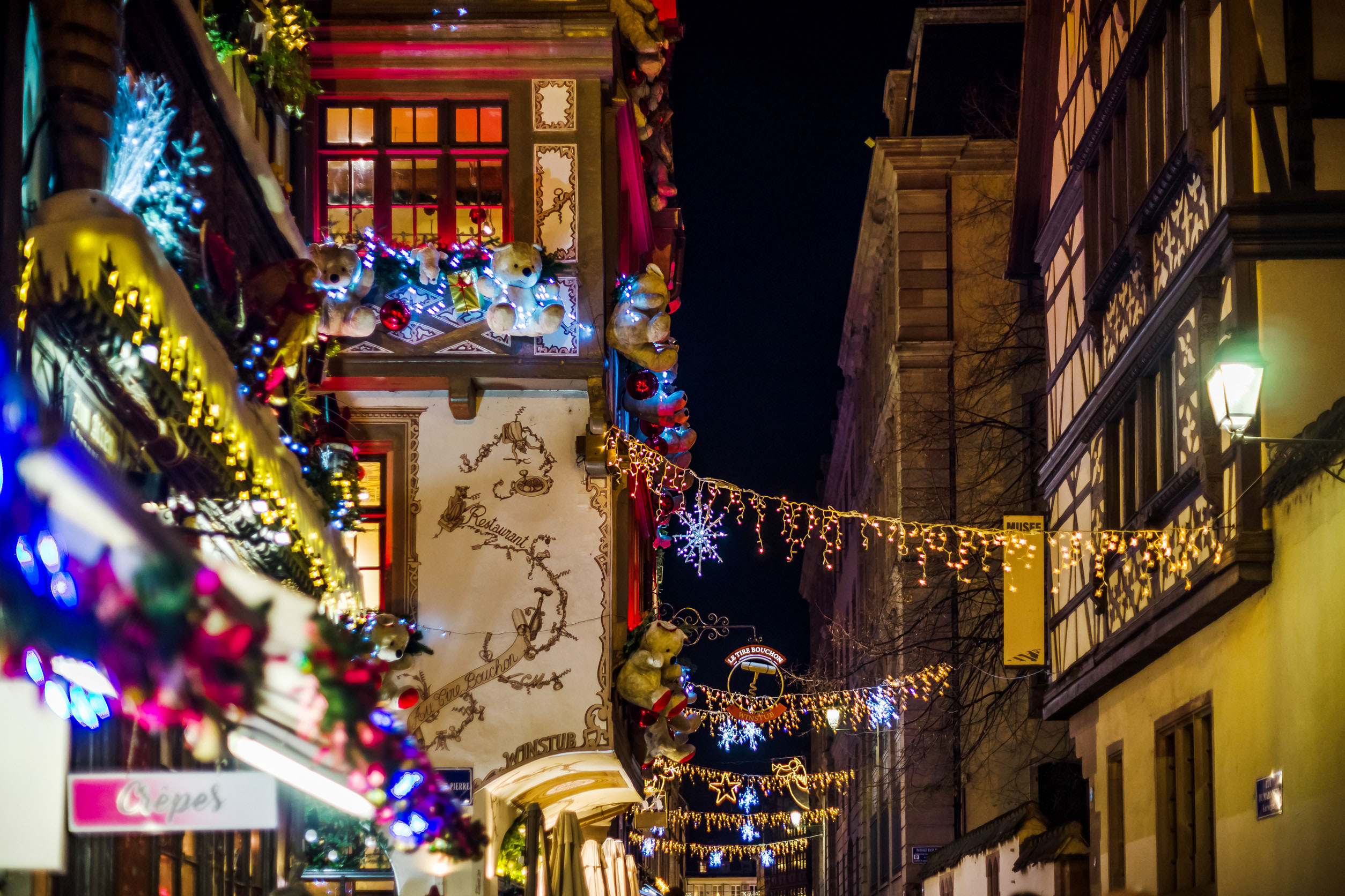“A narrow street in Strasbourg decorated with Christmas lights, ornaments, and festive shopfront displays at night.