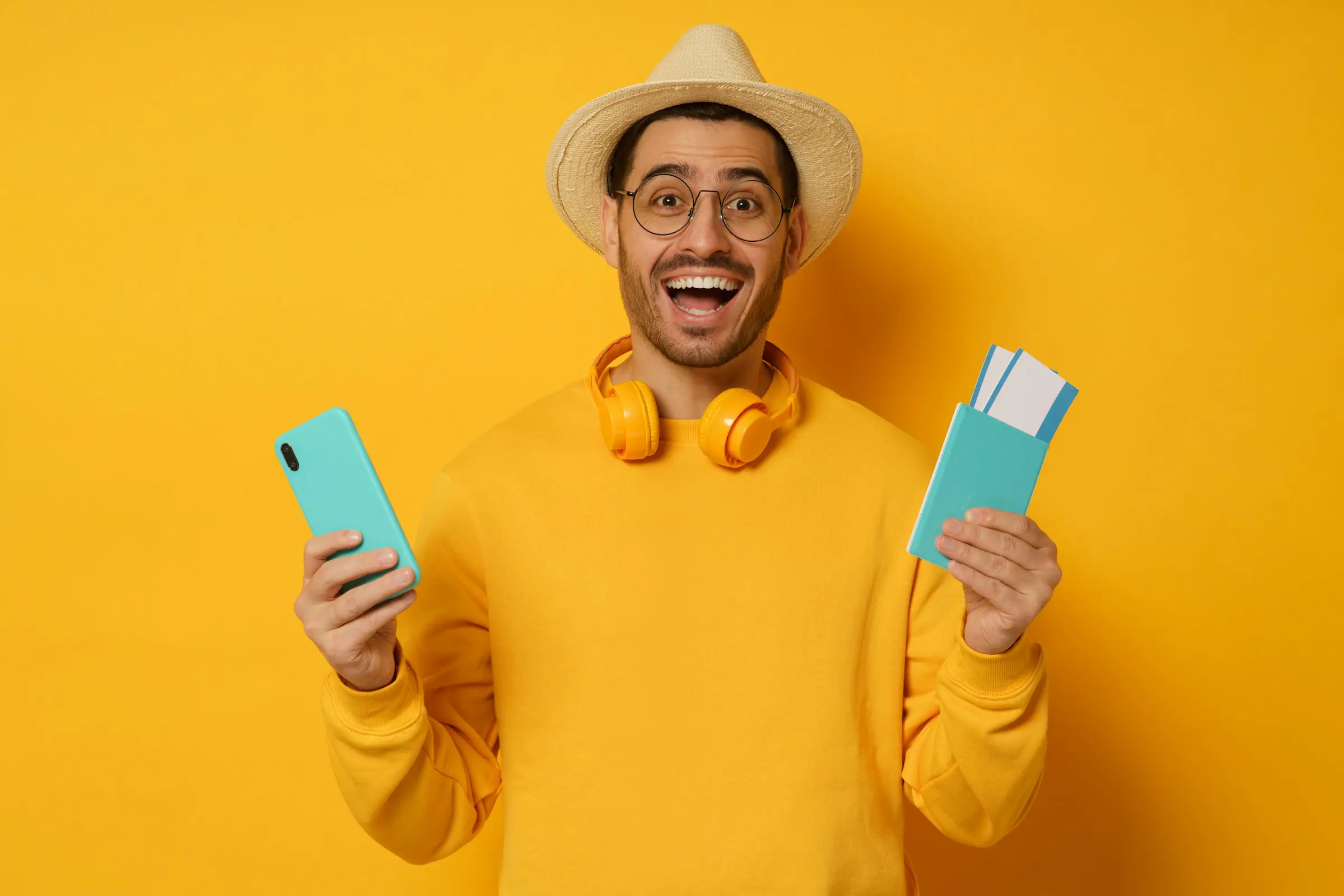 Happy traveler in a yellow sweater and hat holding a smartphone and passport with tickets against a bright yellow background.