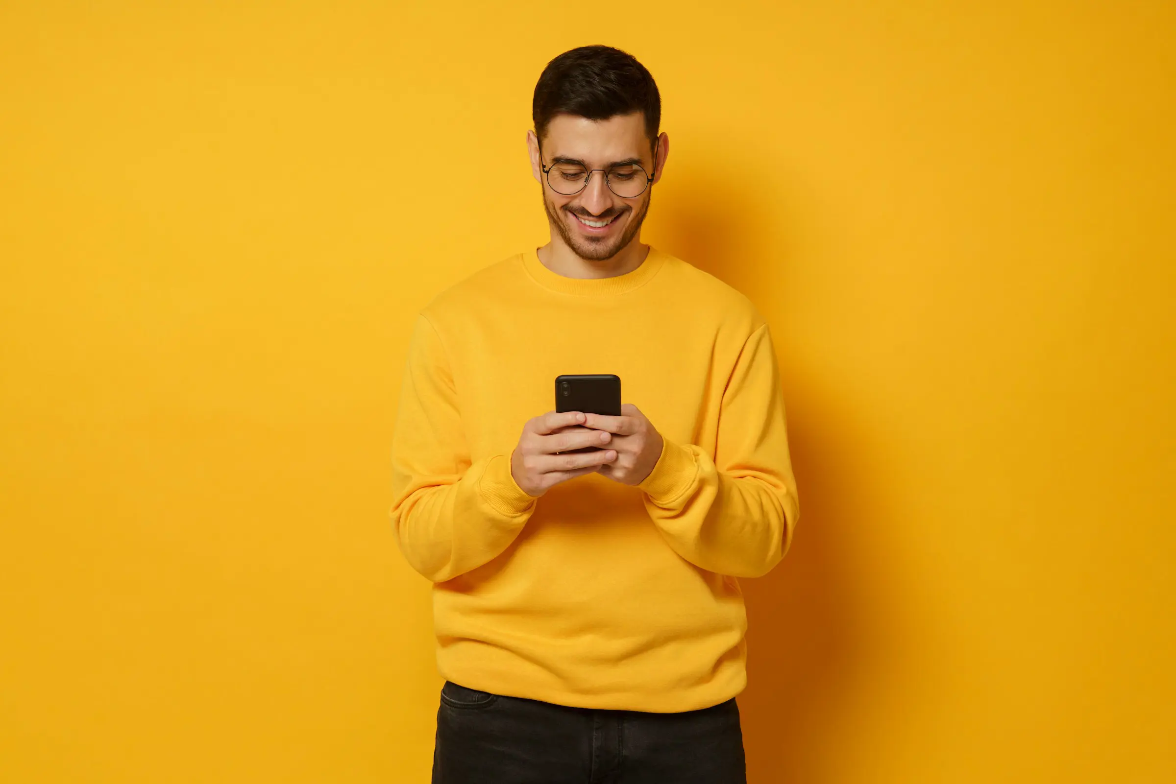 Man in a yellow sweater smiling while using a smartphone against a yellow background.