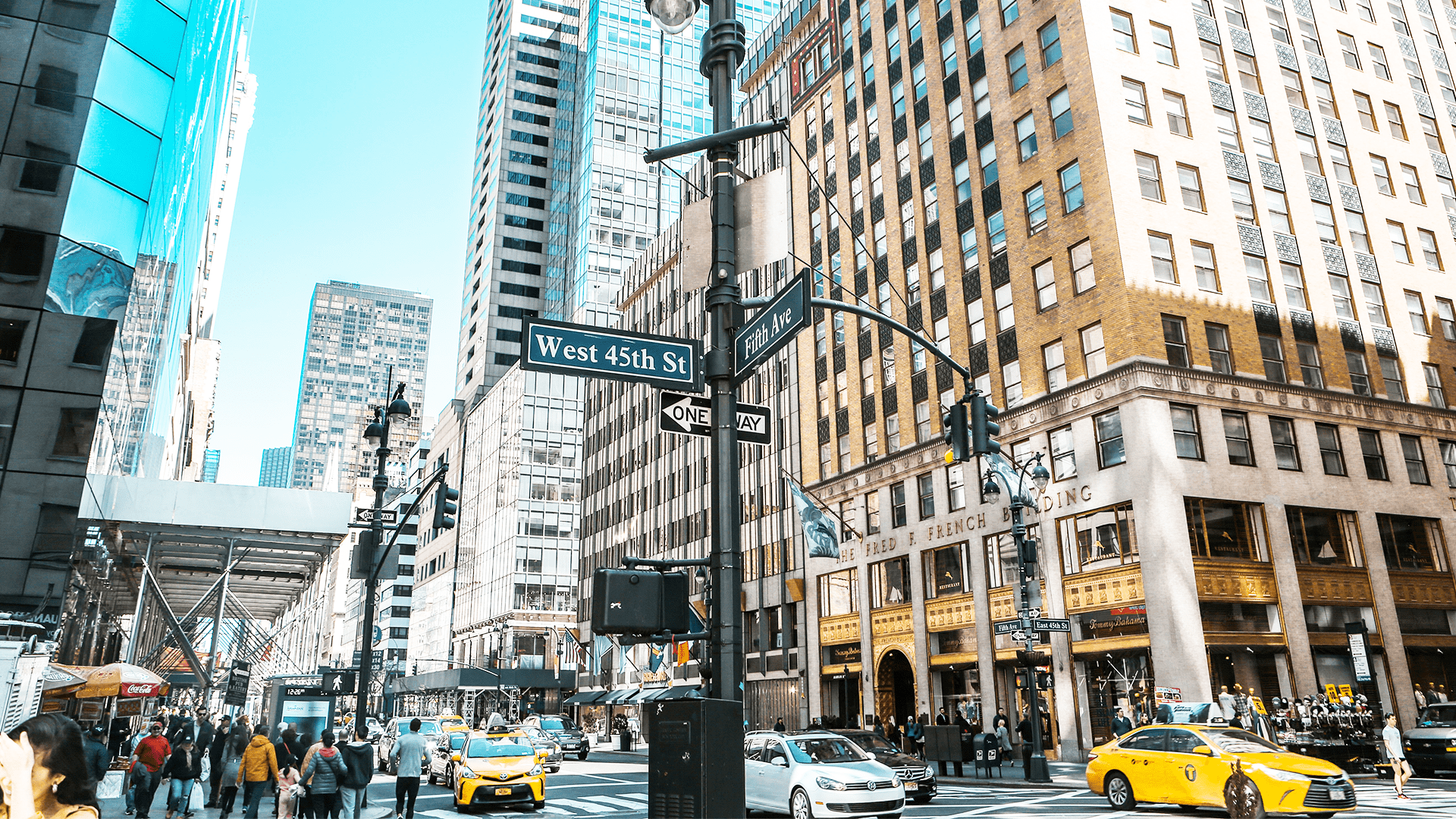 Gatekryss ved West 45th St og Fifth Ave i New York City, med høye bygninger, folk som spaserer, og gule drosjer i trafikken.