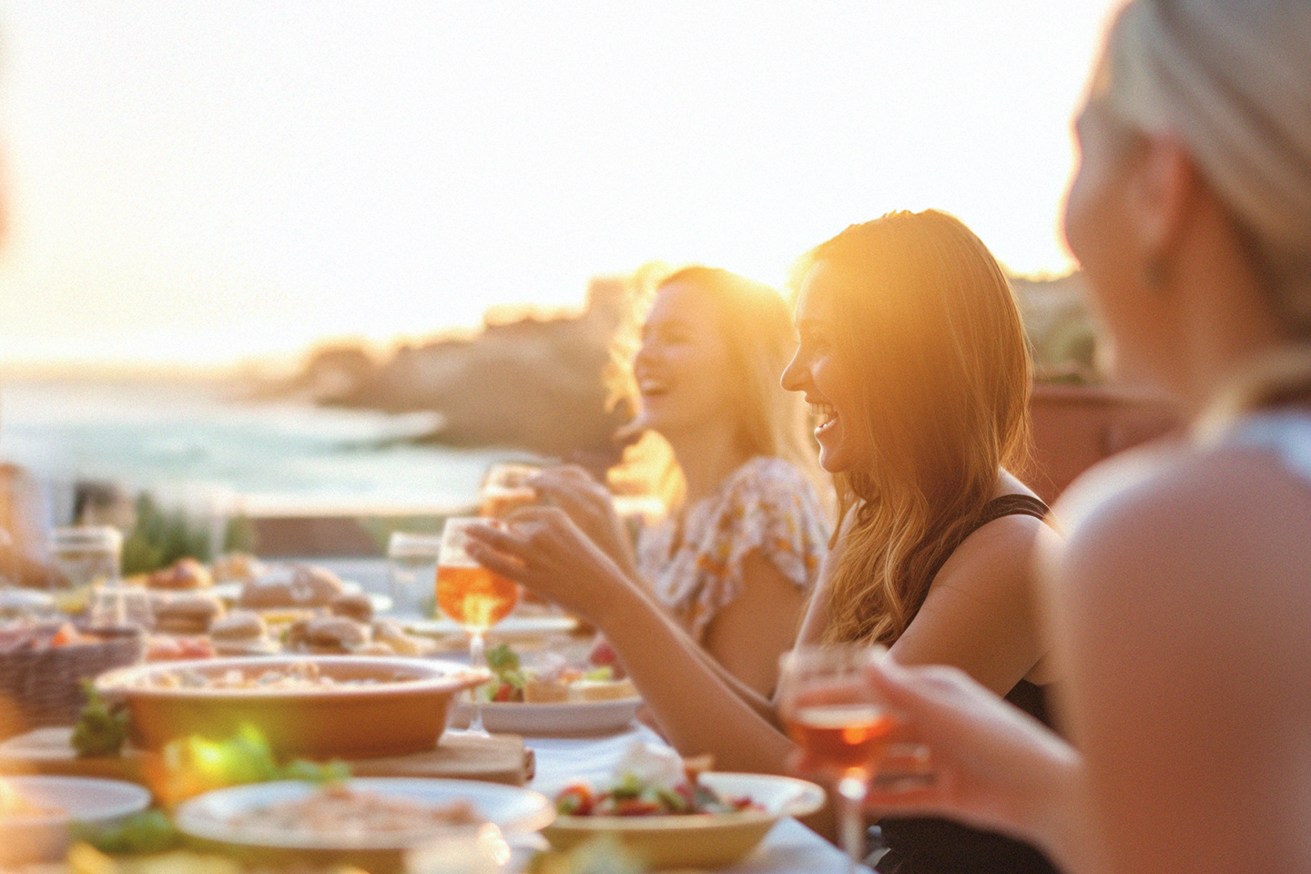Folk som nyter en middag utendørs ved solnedgang, ler og holder glass med drikke.