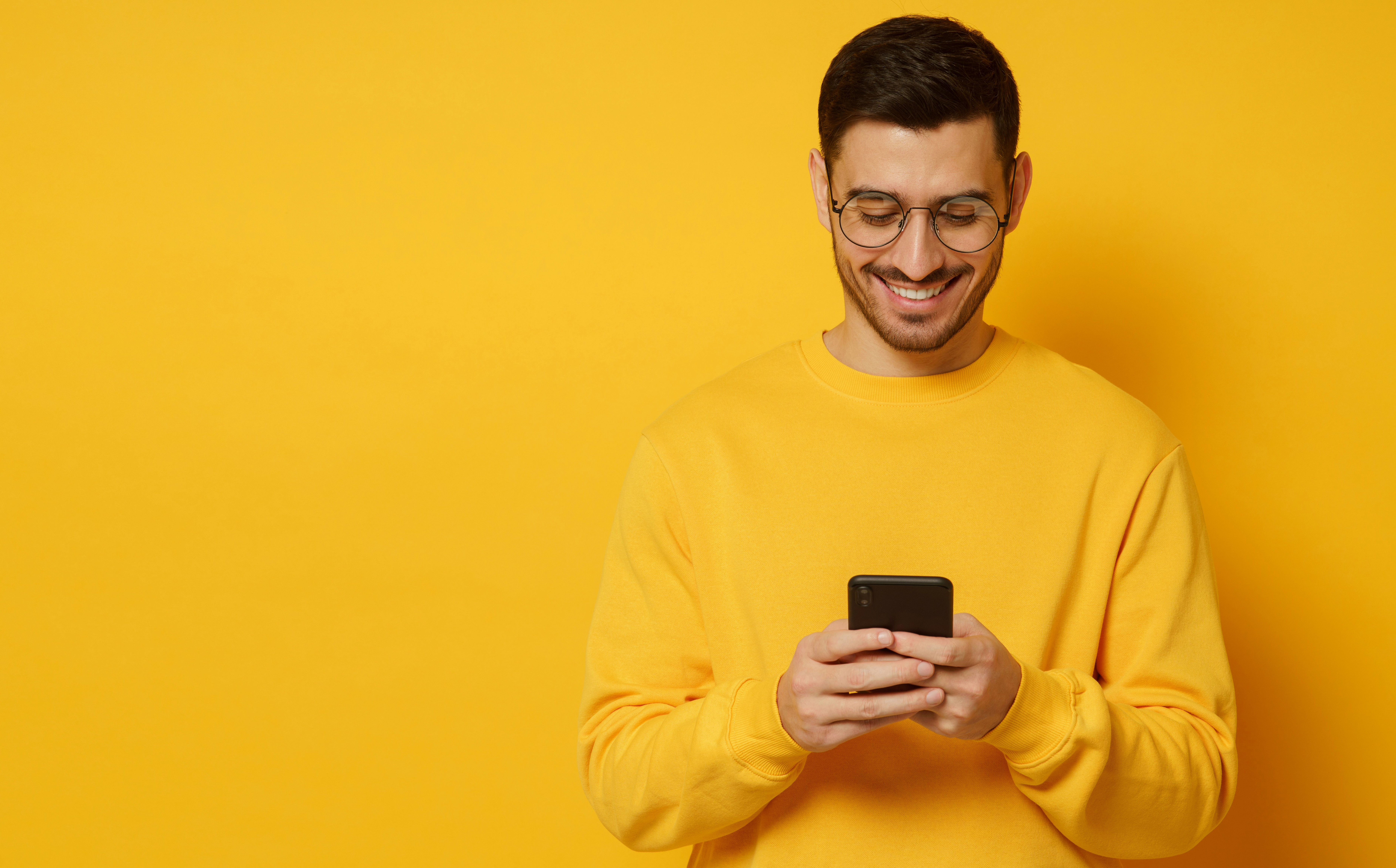 Smiling man in a yellow sweater using a smartphone against a yellow background.
