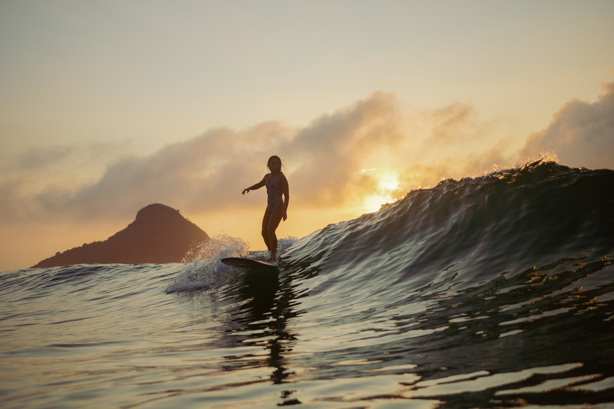 Surfer riding a wave during sunset with a mountain silhouette in the background.