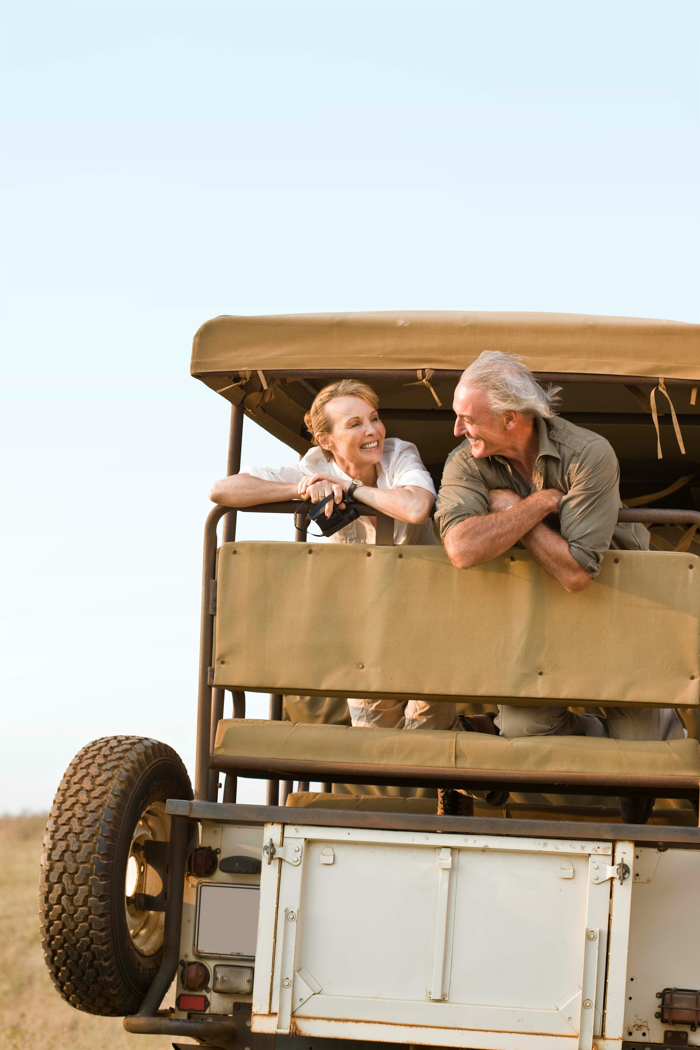 Elderly couple enjoying a safari ride in an open jeep, smiling and leaning on the vehicle's backrest, with a clear sky in the background.