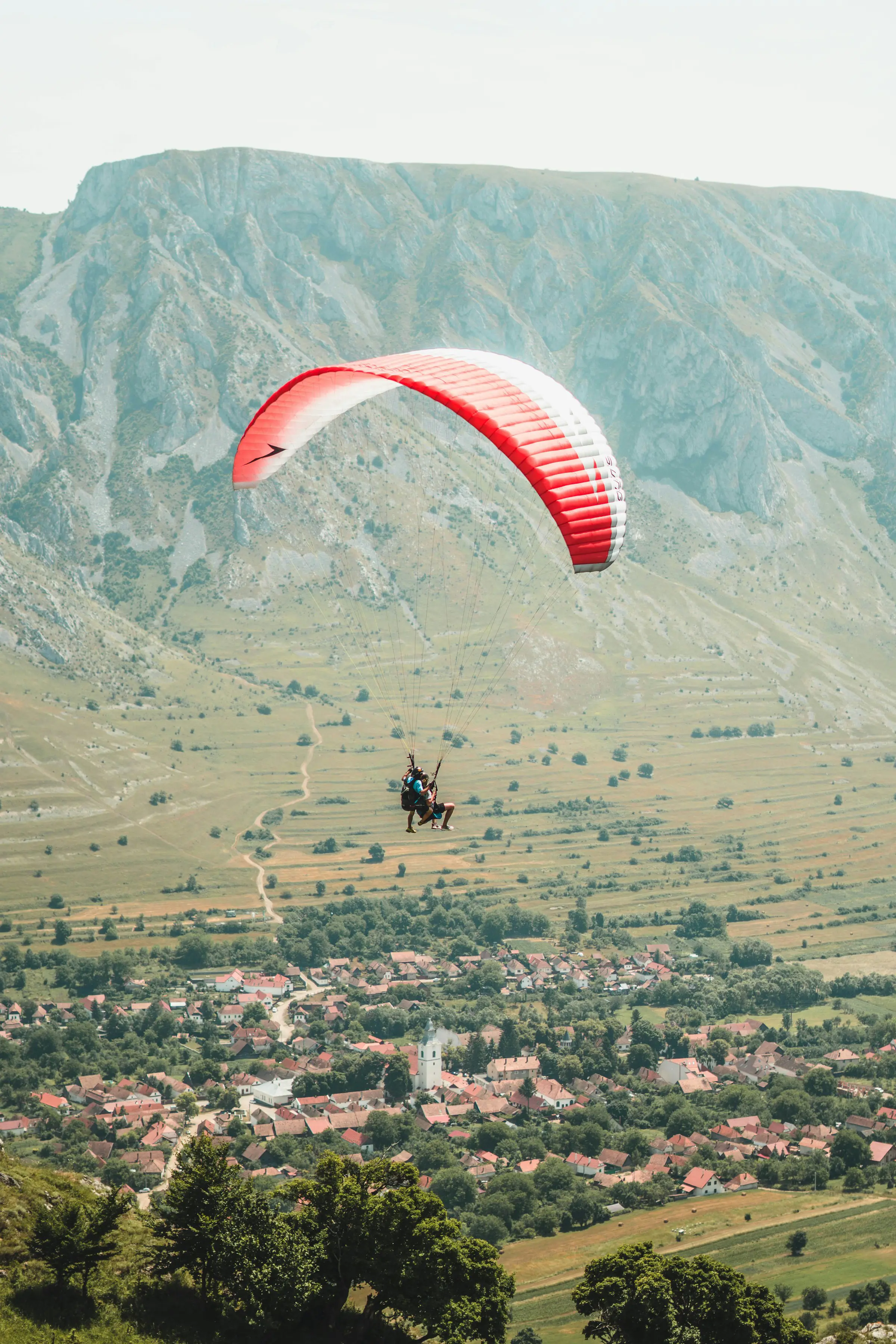 Paraglider flying over a scenic rural landscape with a small village and rolling hills below.