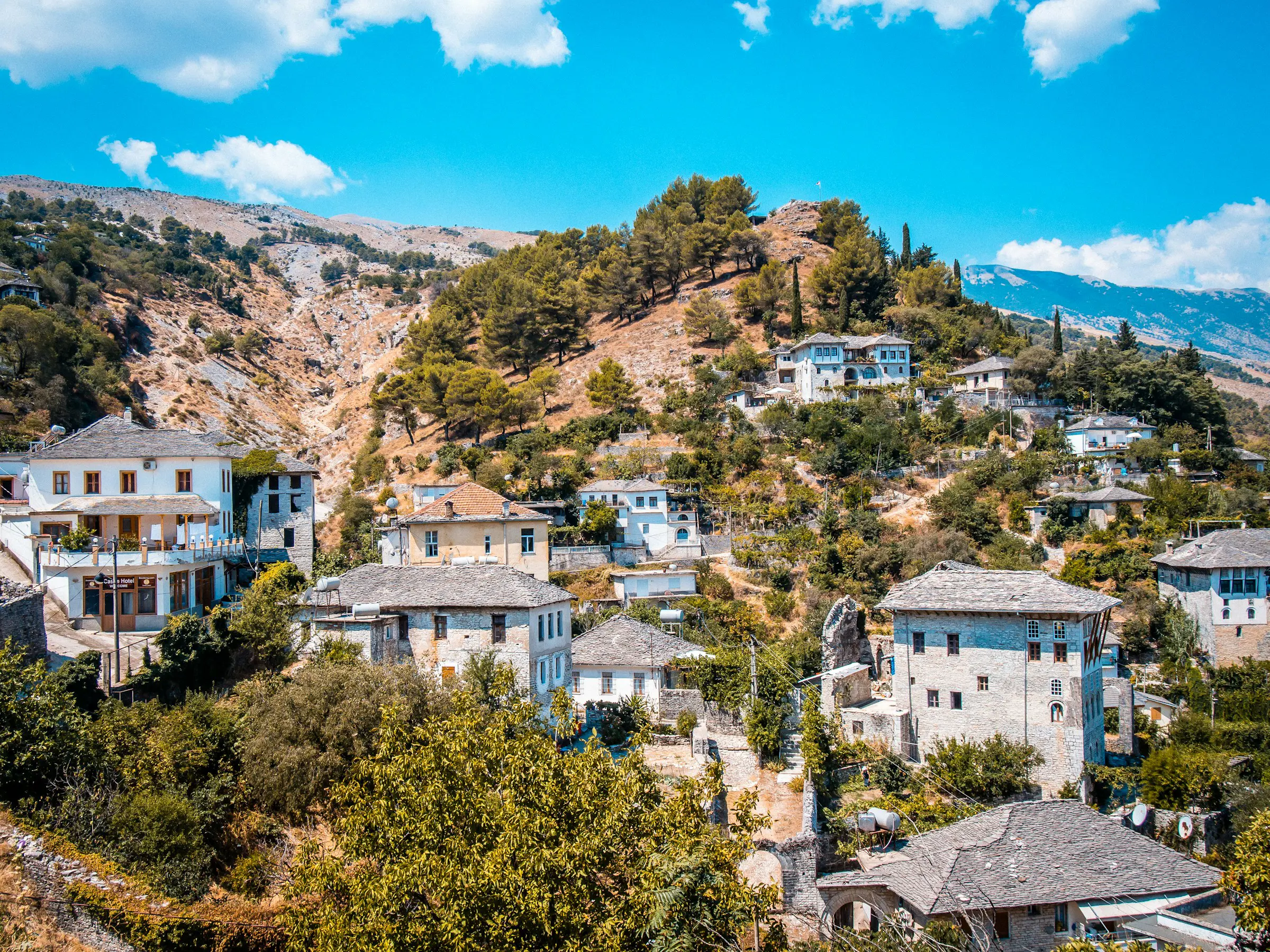 Scenic view of a traditional hillside village in Albania with stone houses and lush greenery under a bright blue sky.