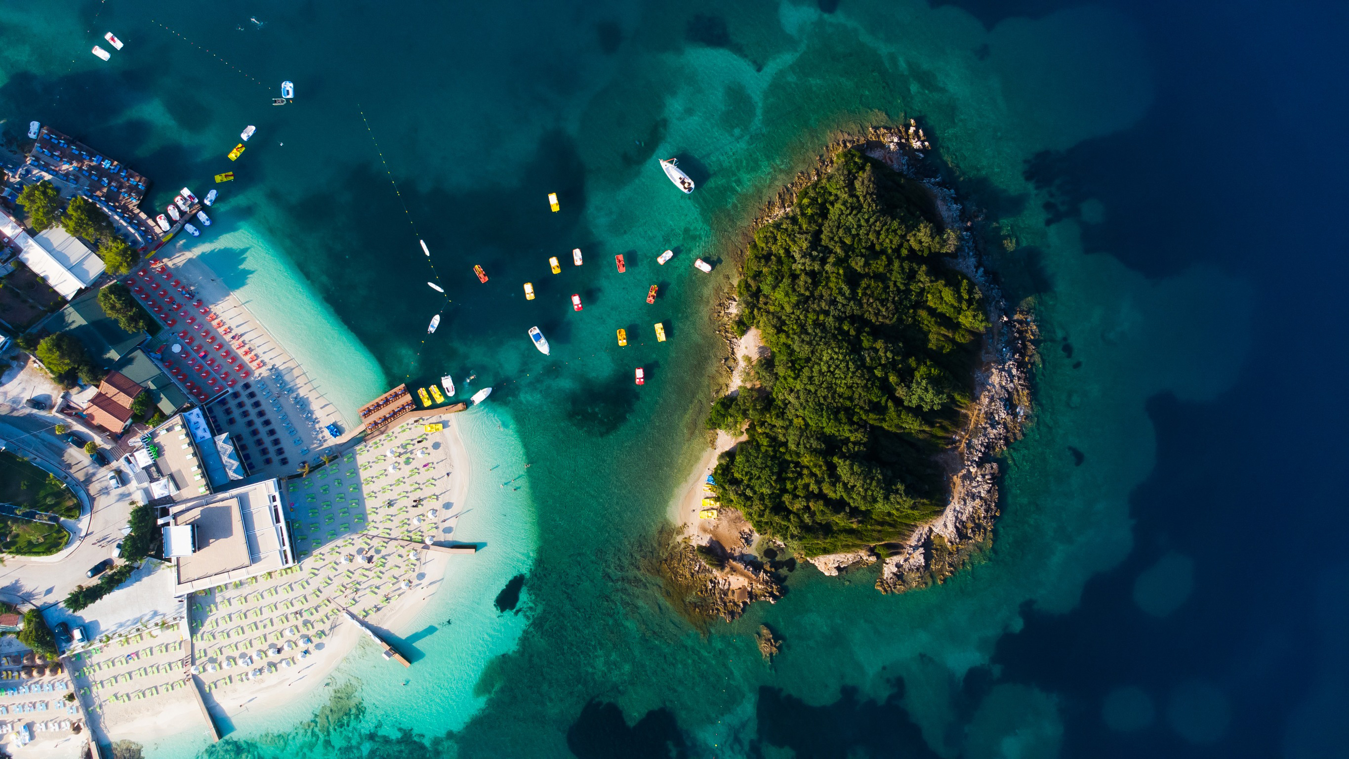 Travel to Saranda-Ksamil - View of a tropical island with lush greenery surrounded by clear turquoise water and sandy beaches, boats anchored nearby.
