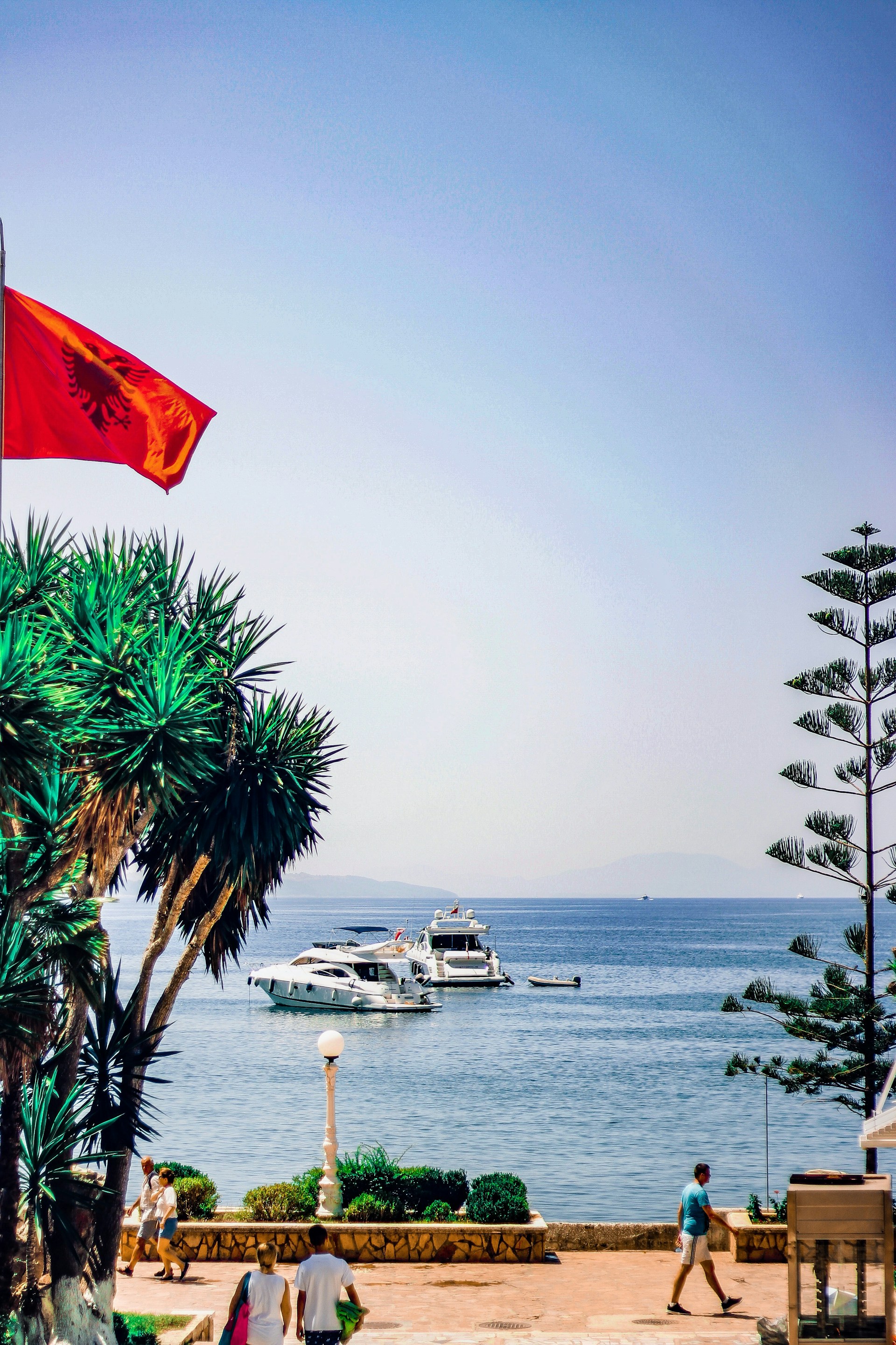 Kystutsikt i Albania med rødt flagg, to båter i klart blått hav og palmetrær, med folk som går langs promenaden