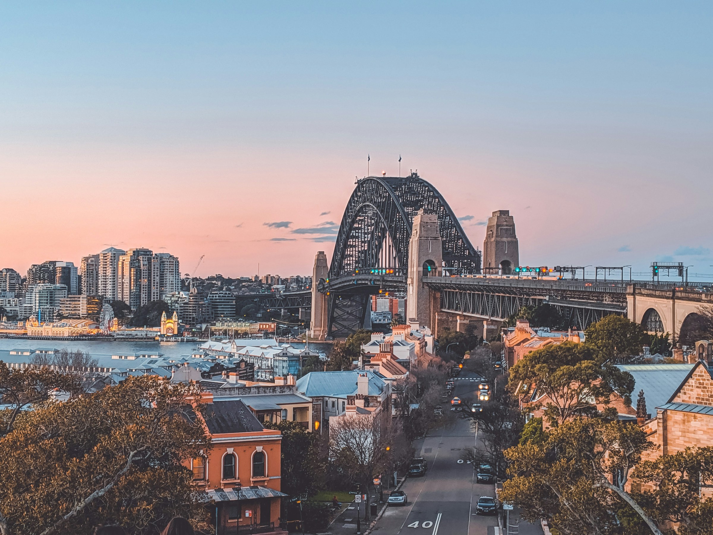 Scenic view of Sydney Harbour Bridge at sunset with urban skyline and waterfront buildings.