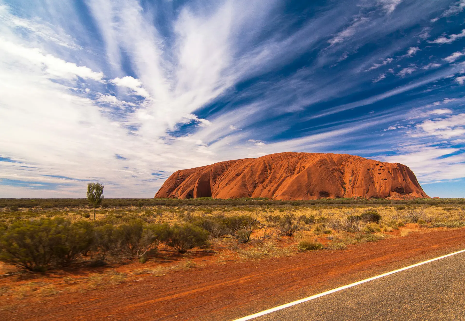 Uluru, en stor rød sandsteinsmonolitt, omgitt av tørt landskap under en blå himmel med skyer i Australia