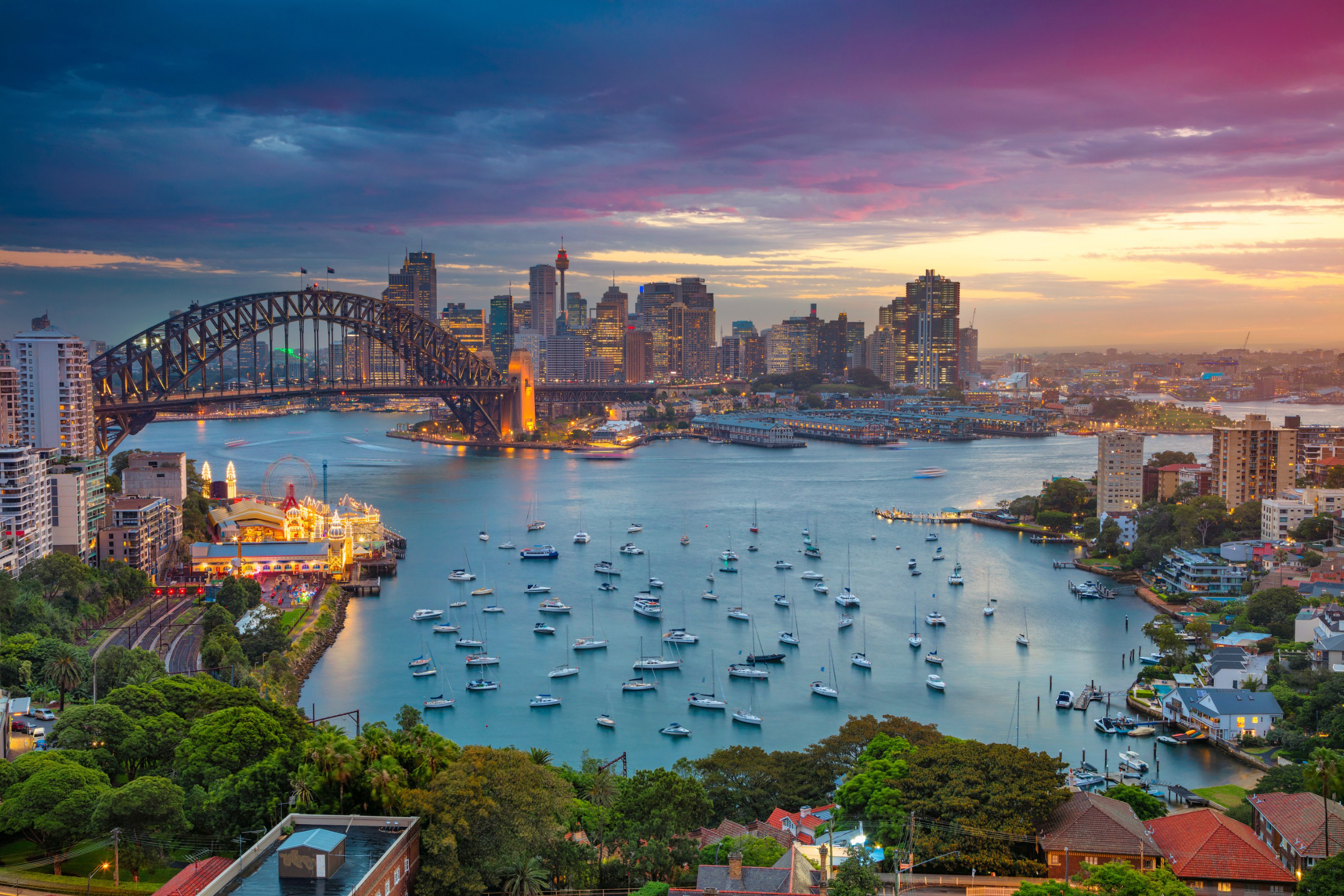 Travel to Sydney - Sydney harbor skyline at sunset with the Sydney Harbour Bridge, city buildings, and boats on the water.