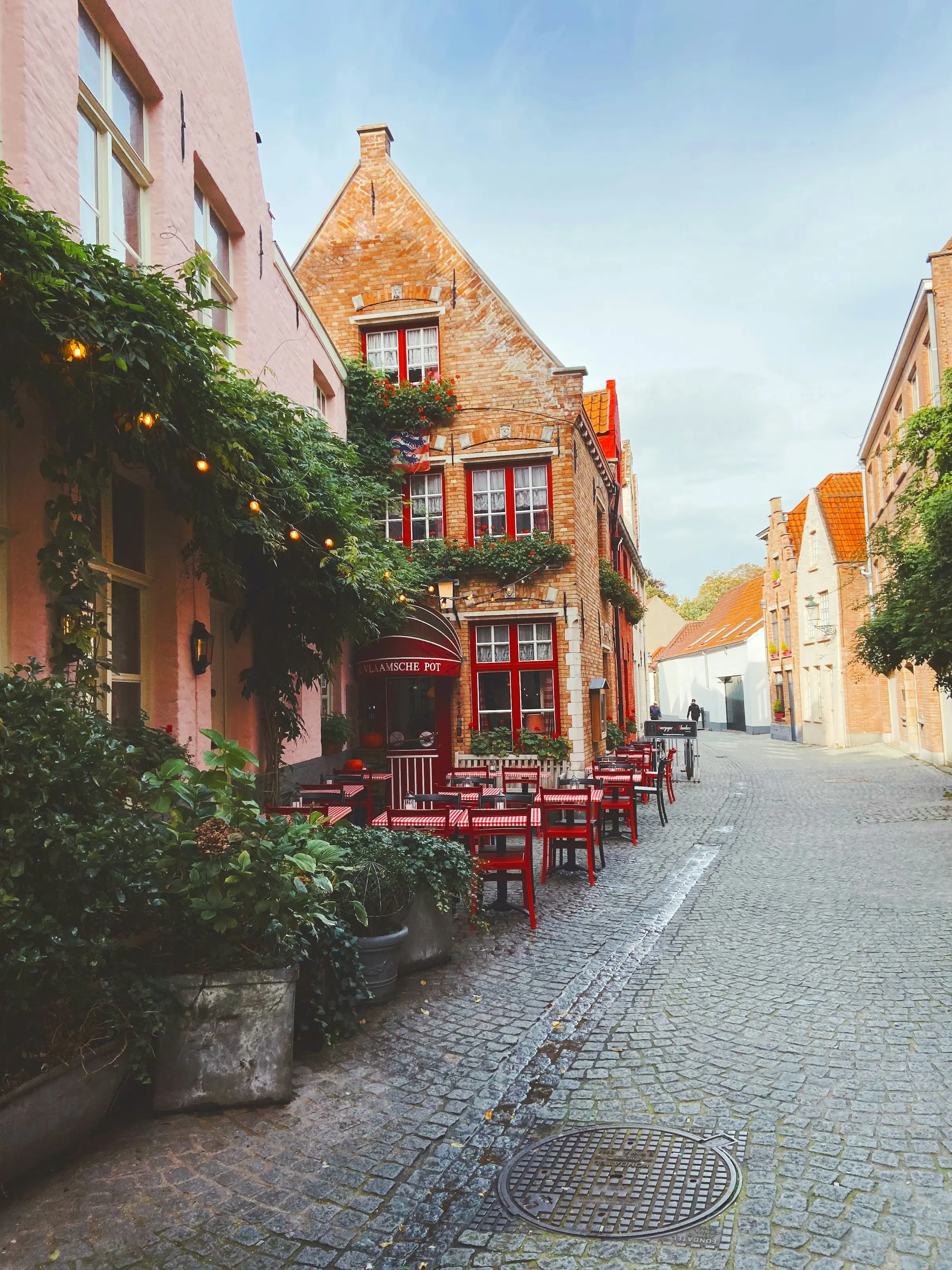 Charming European cobblestone street with historic brick buildings, ivy-covered walls, and a quaint outdoor cafe with red chairs and tables.