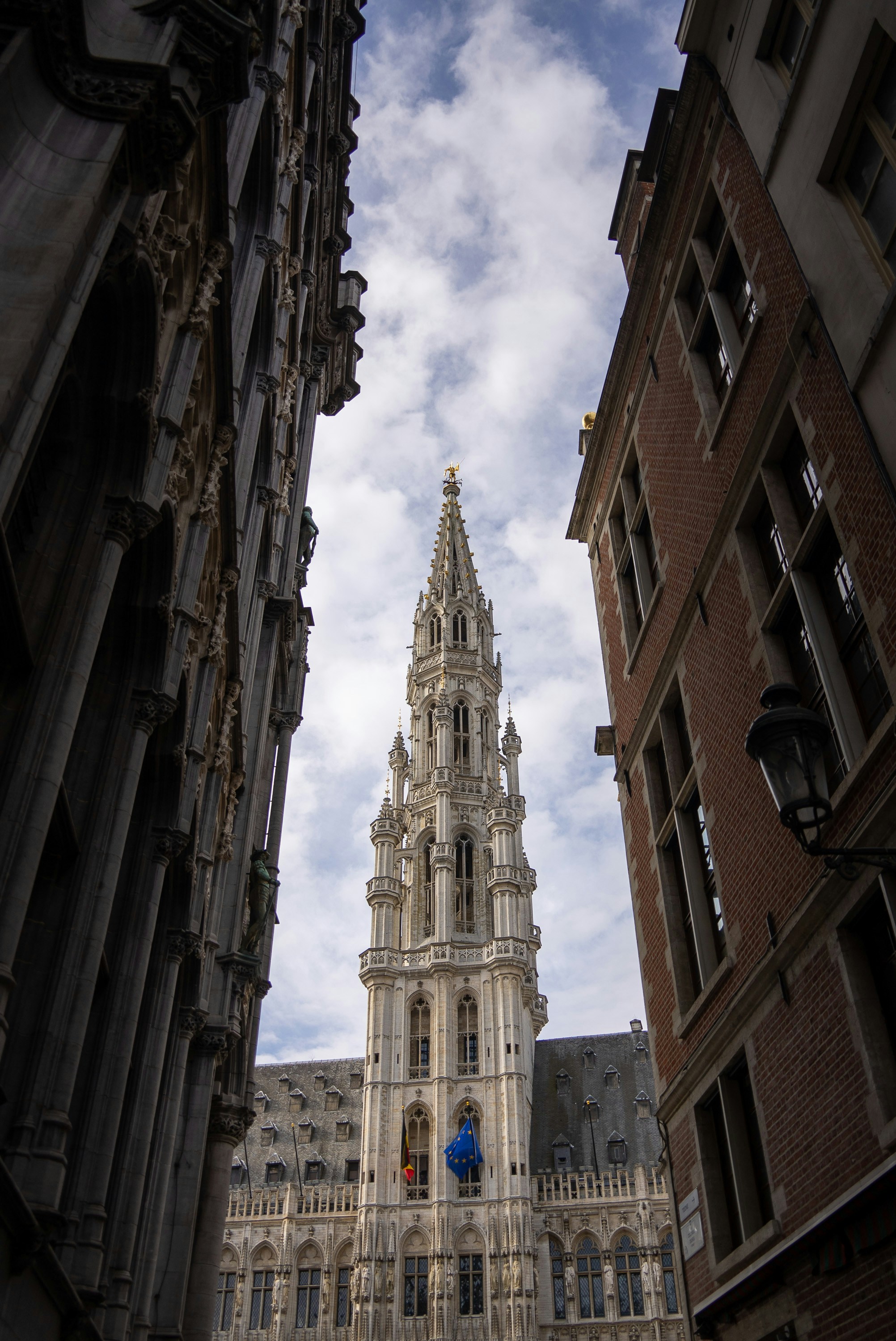 Rådhustårnet på Grand Place i Brussel, sett mellom to bygninger, under en delvis skyet himmel. Belgisk og EU-flagg vaier