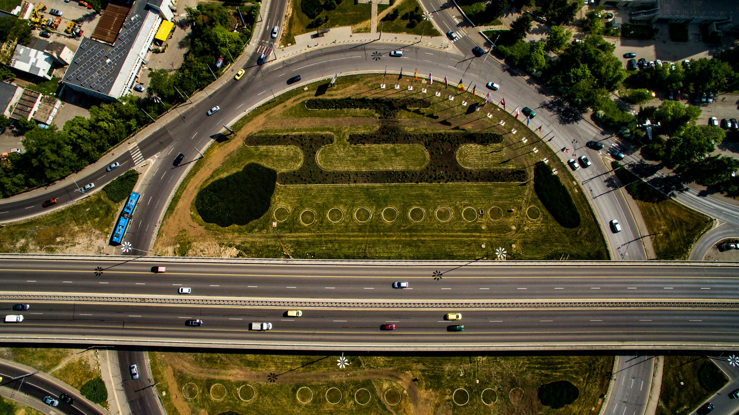 Aerial view of a green roundabout with creative landscaping design, surrounded by busy roads and vehicles, highlighting urban planning and infrastructure integration.