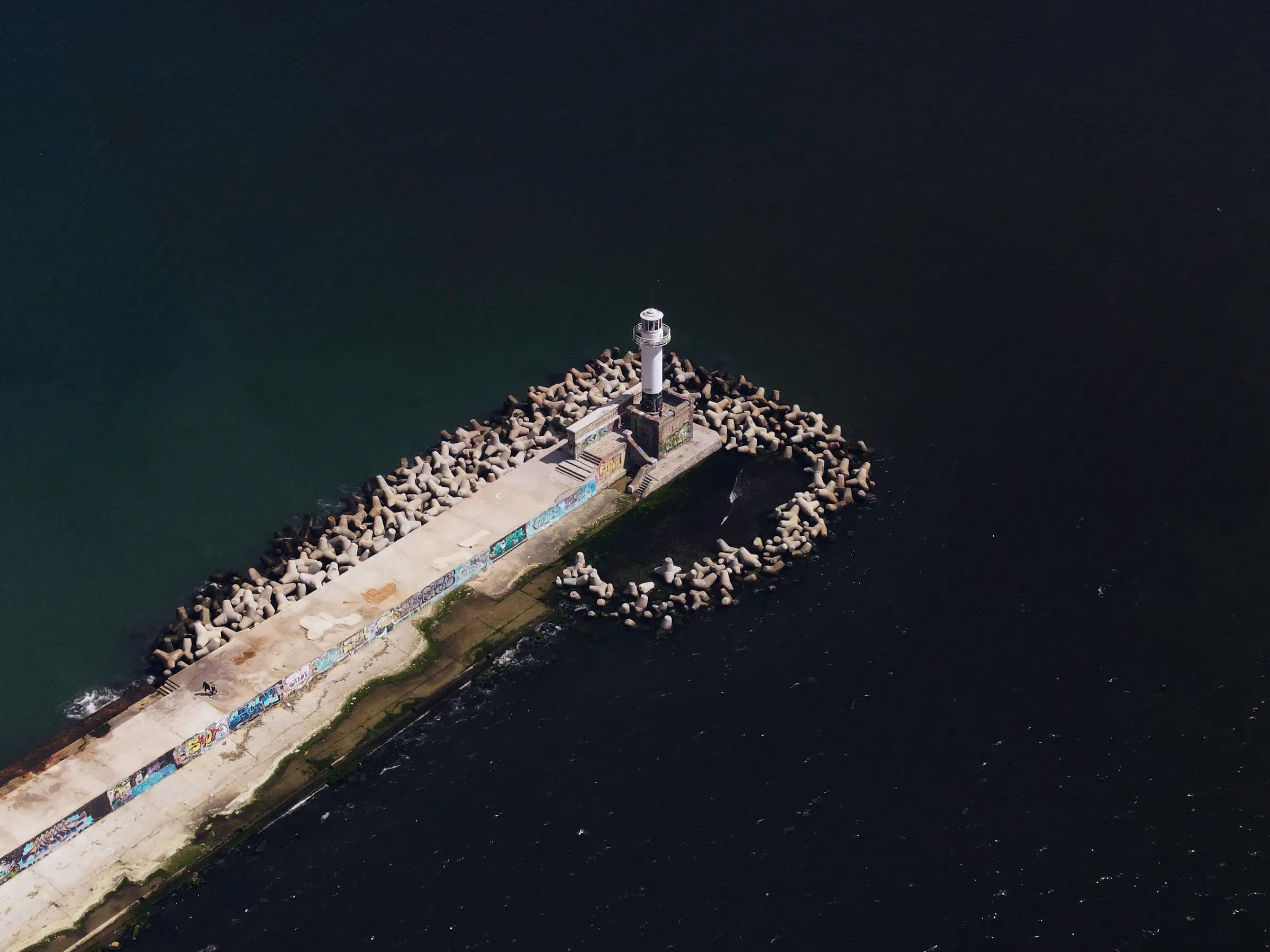 Aerial view of a lighthouse on a concrete pier surrounded by wave breakers extending into dark waters, with graffiti on the pier's surface.
