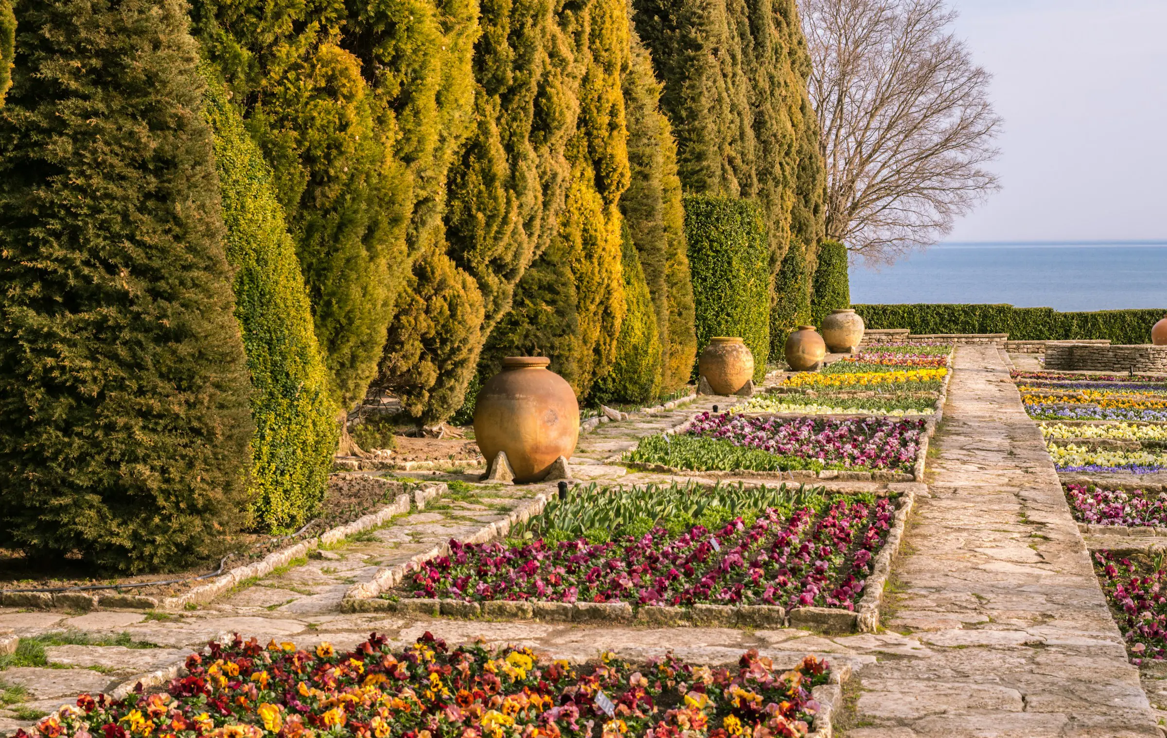 Colorful flower garden with stone paths, ceramic pots, and lush greenery, overlooking a calm sea.