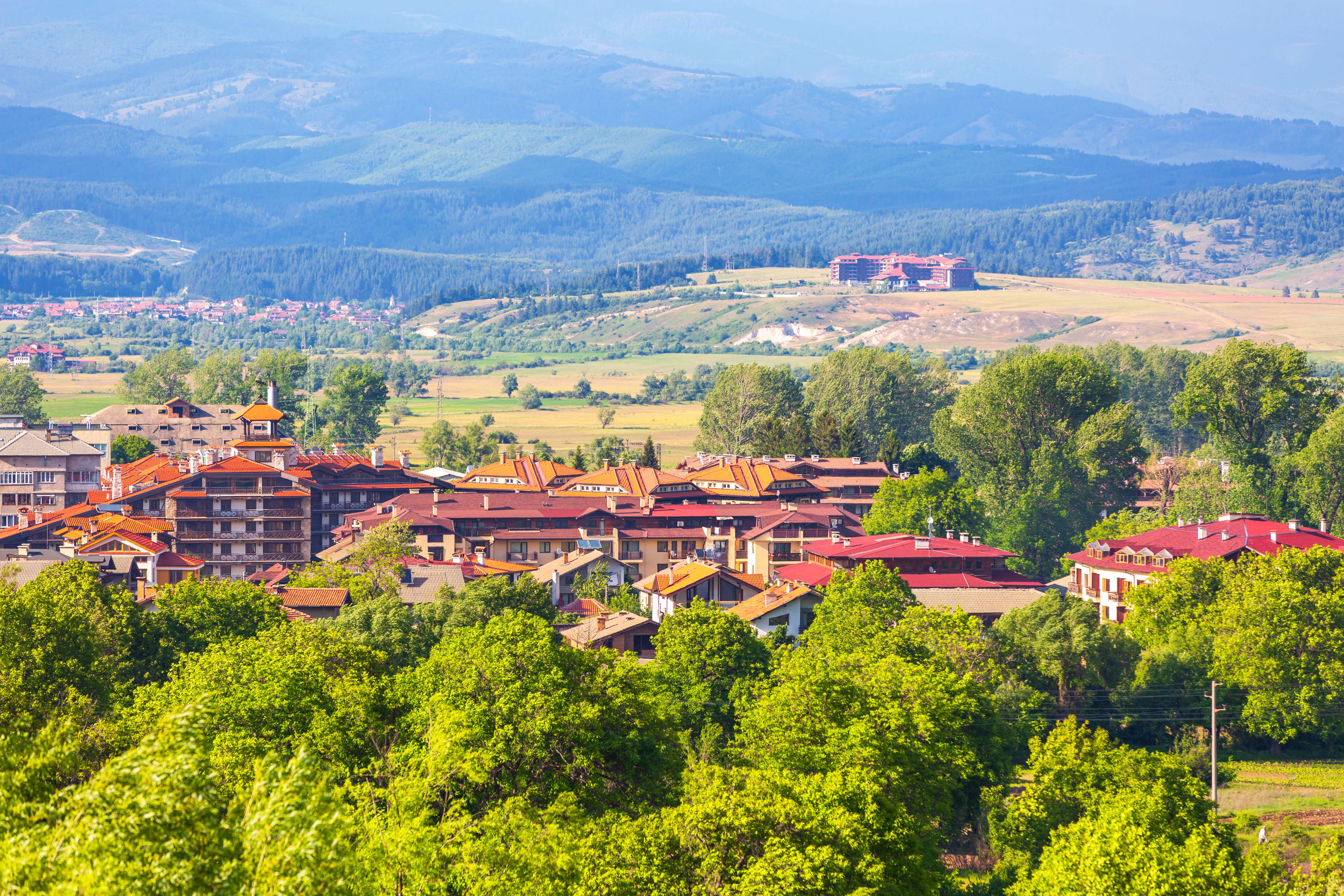 Utsikt over en landsby omgitt av frodig landskap og skogkledde åser i bakgrunnen, med typiske hus med rødbrune tak