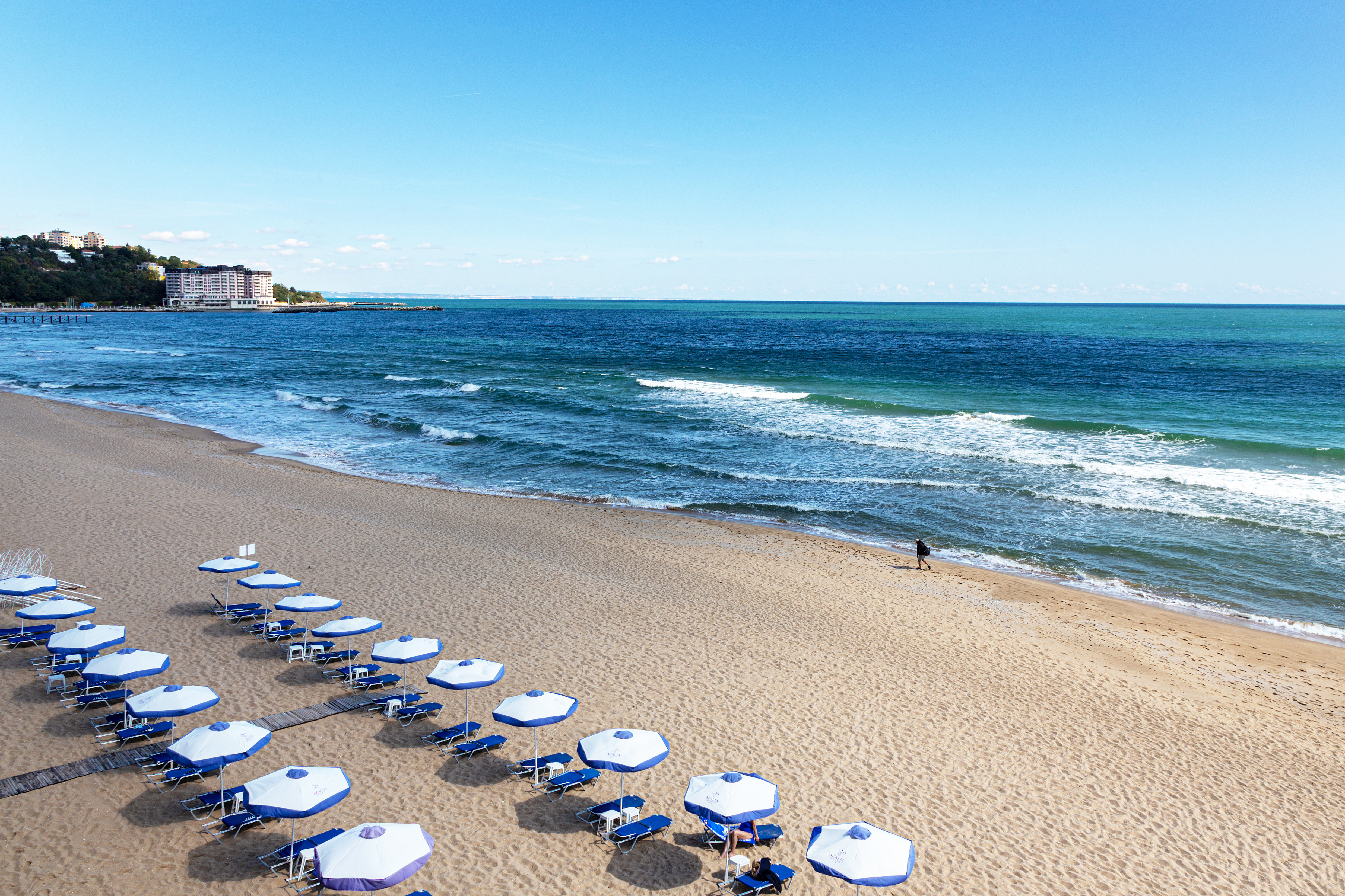 Travel to Sunny Beach - Sandy beach with rows of blue and white umbrellas facing the ocean waves under a clear blue sky.