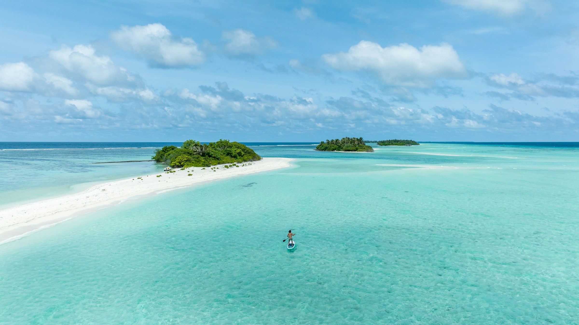 A person paddleboarding in clear turquoise water near small tropical islands with lush greenery under a blue sky.