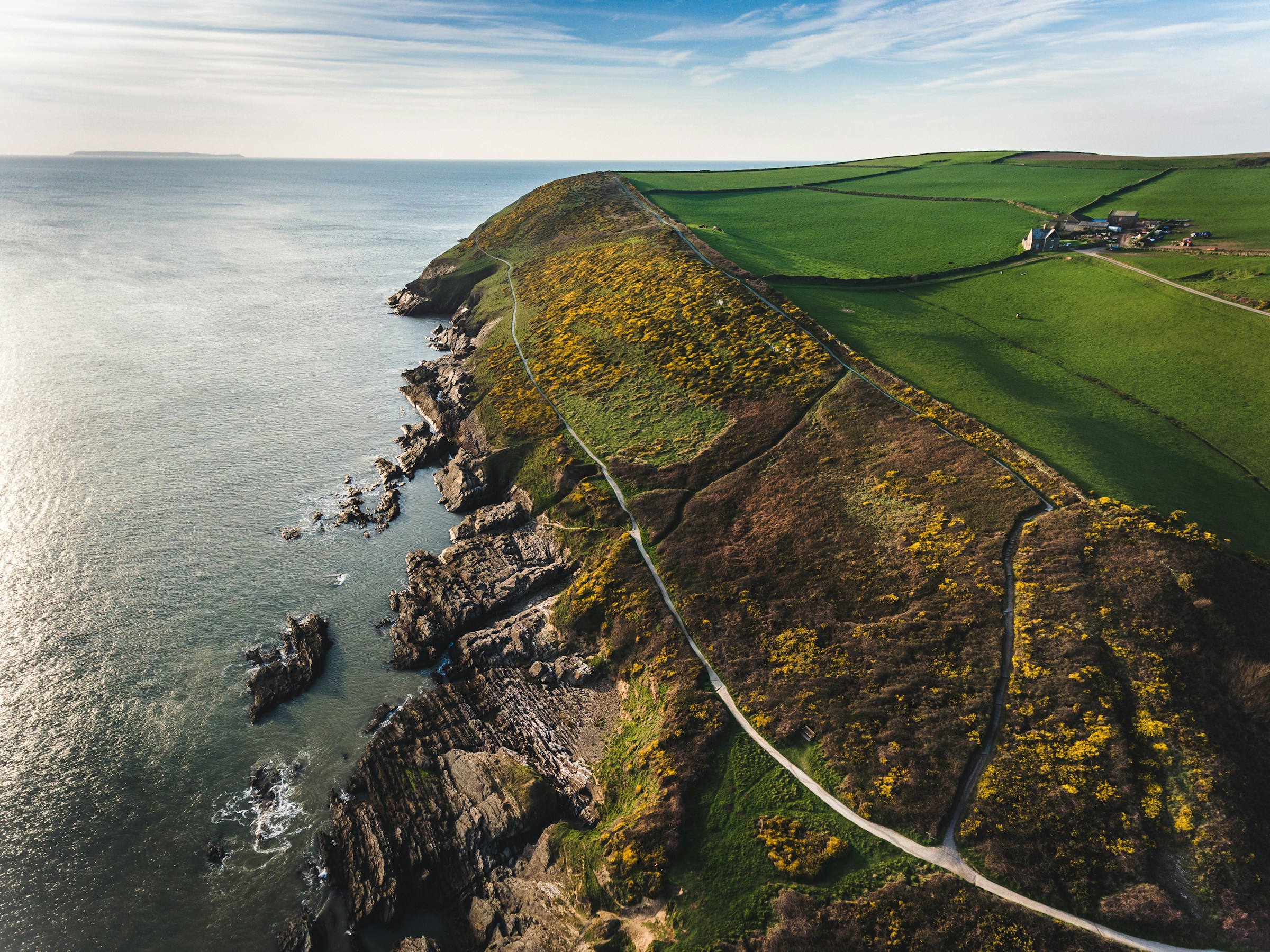 Aerial view of a coastal cliff landscape with green fields, a winding path, and rocky shoreline beside the calm ocean under a clear blue sky.