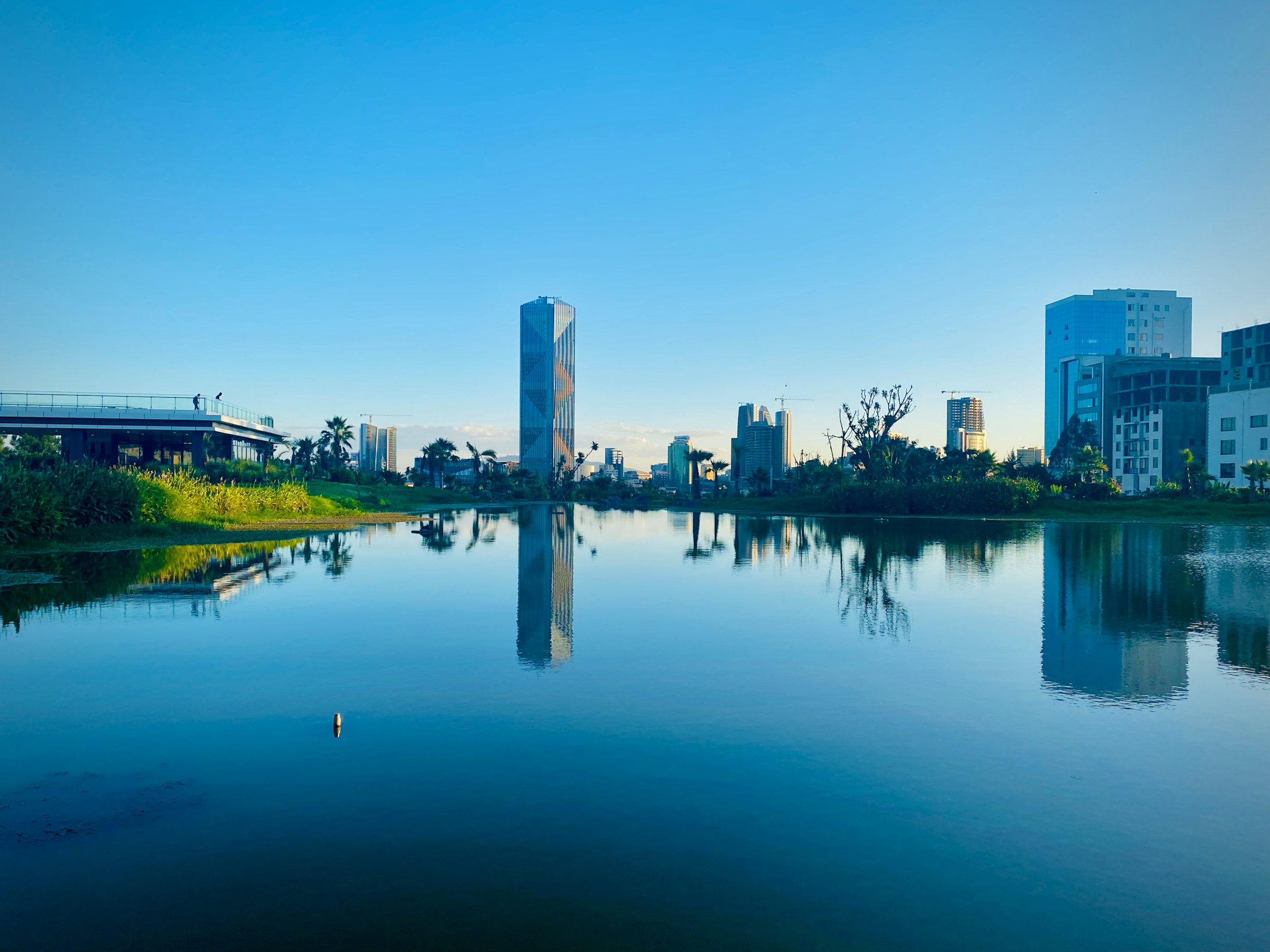 Modern city skyline reflected in a serene lake under a clear blue sky.