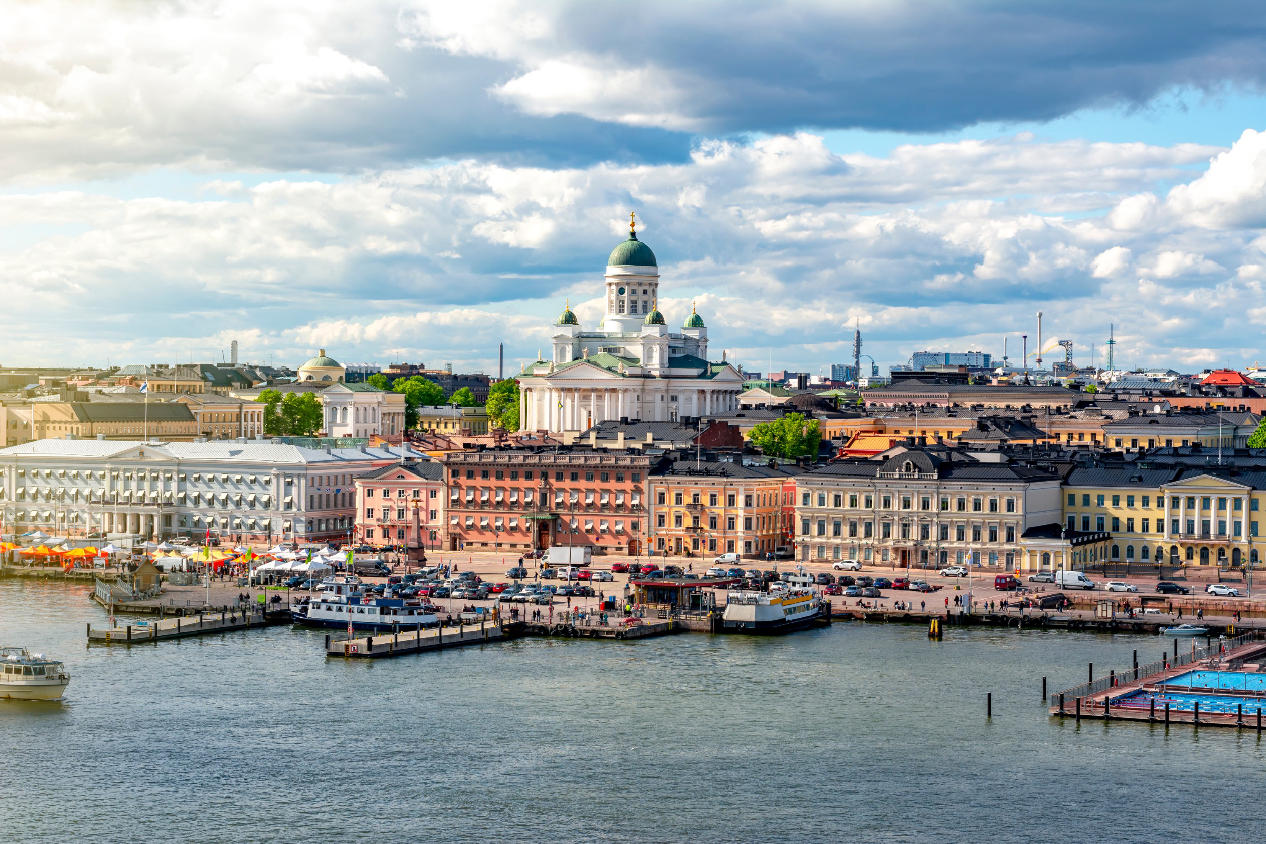 Helsinki-bybilde med Uspenskijkatedralen i bakgrunnen, fargerike bygninger ved vannkanten, og båter i havnen under en delvis skyet himmel
