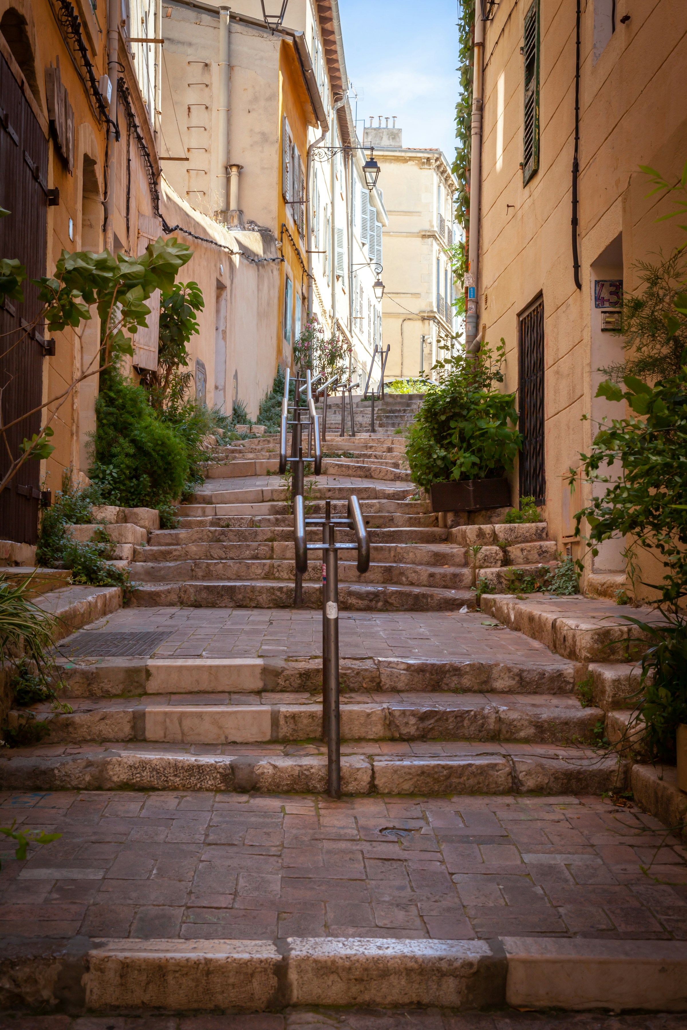 Narrow European alleyway with stone steps and handrails, surrounded by rustic buildings and greenery under a clear sky.
