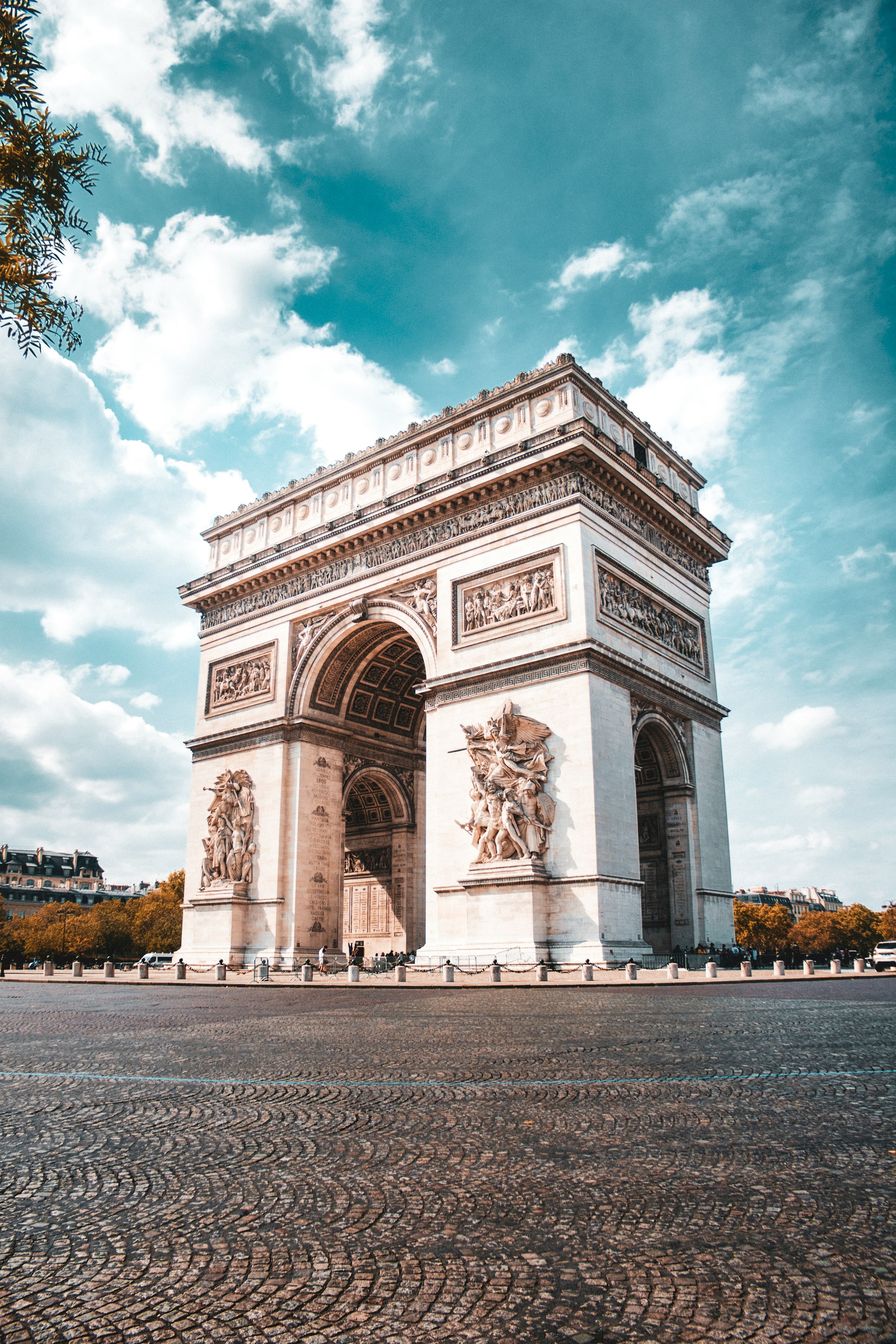 Arc de Triomphe in Paris under a partly cloudy sky, surrounded by cobblestone pavement and lined with trees in autumn.