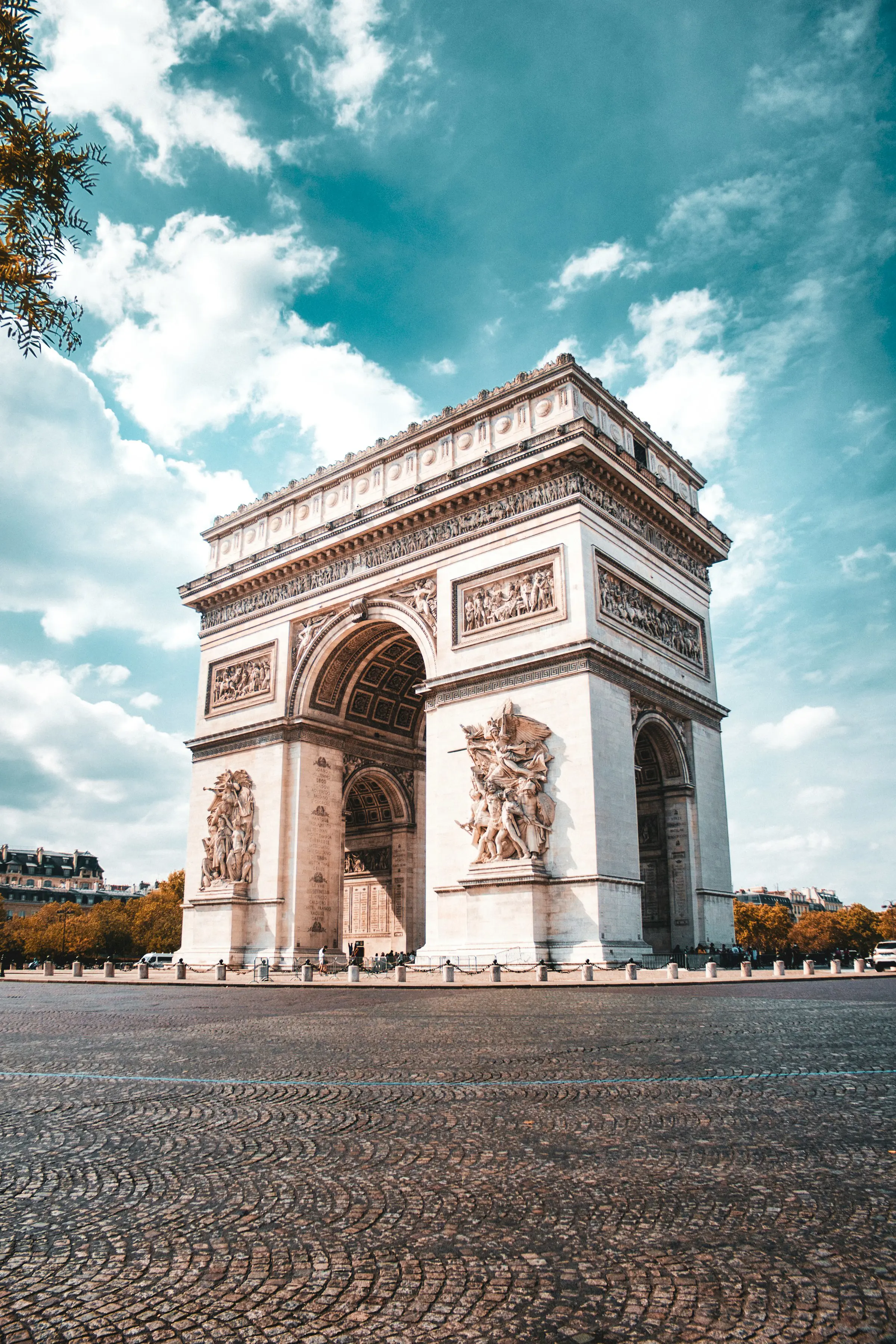 Arc de Triomphe in Paris under a partly cloudy sky, surrounded by cobblestone pavement and lined with trees in autumn.