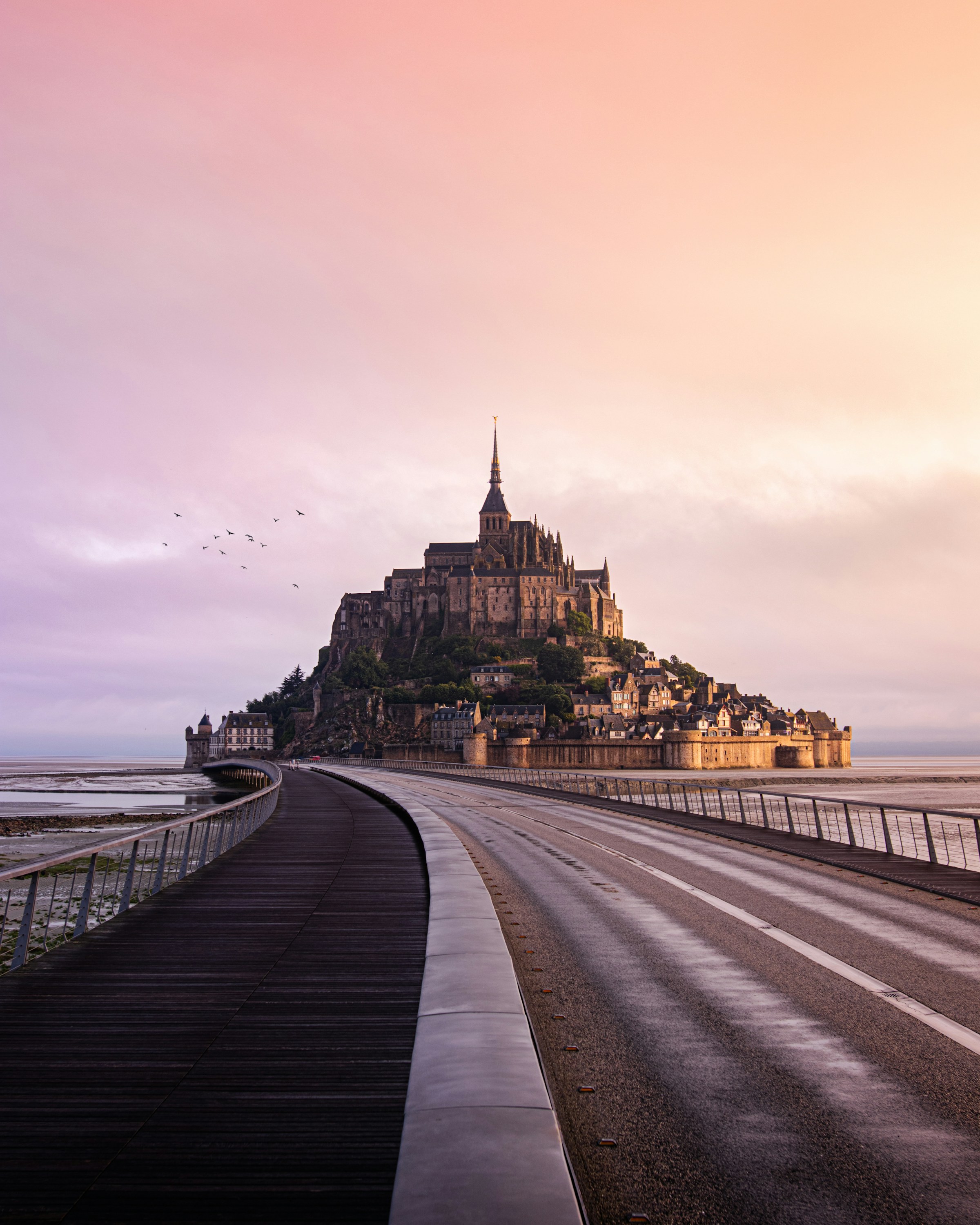 Mont Saint-Michel at sunset with a winding road leading up to the historic island commune, surrounded by the sea. 
