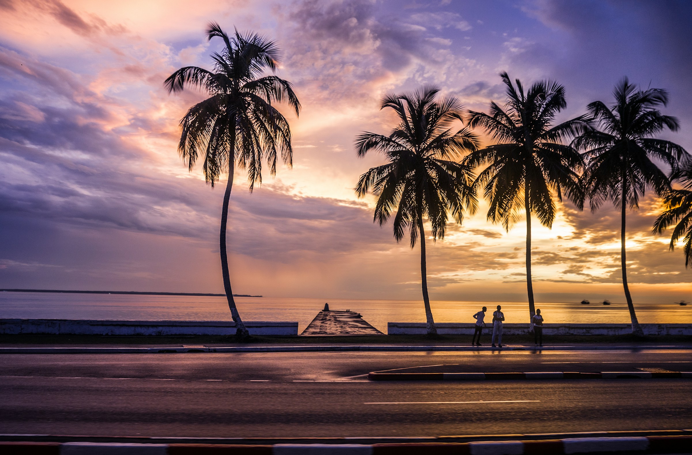 Tropical sunset scene with palm trees silhouetted against a vibrant sky, people walking near the oceanfront, and a picturesque pier leading into the water.