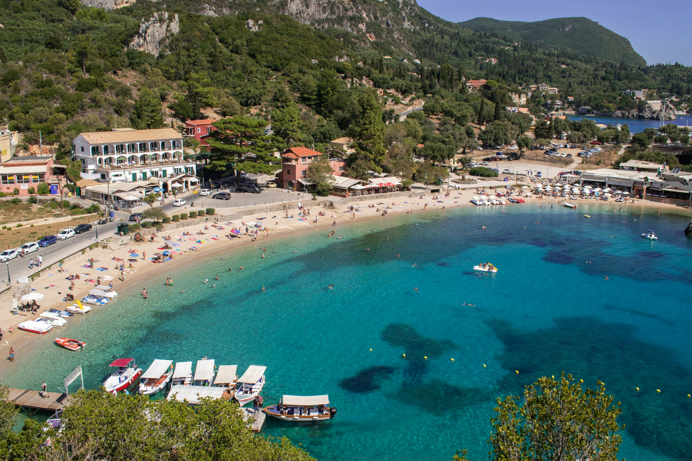 Aerial view of a scenic beach with clear turquoise water, lined with colorful umbrellas, surrounded by lush greenery and hills in the background.