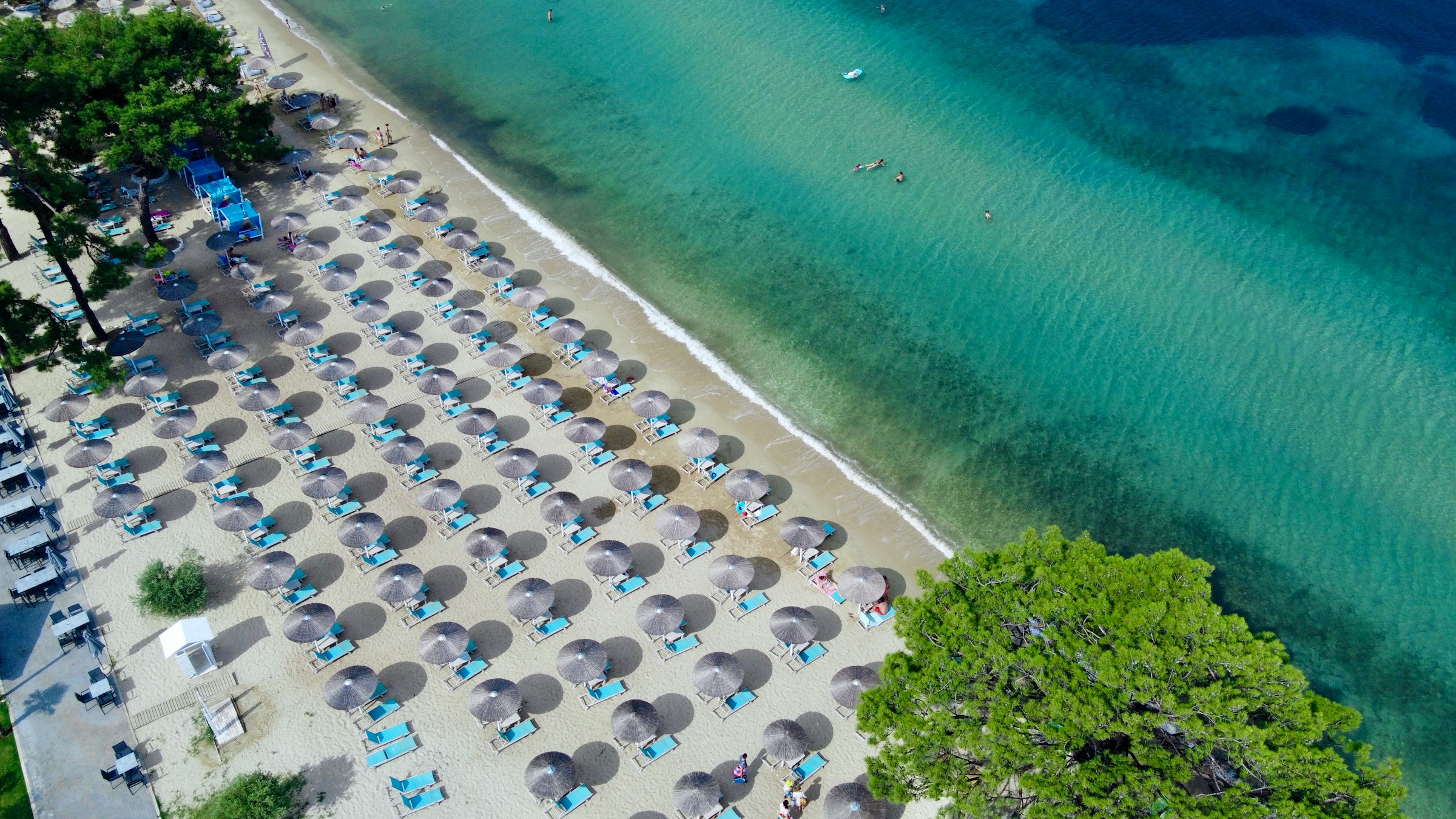 Aerial view of a sandy beach lined with blue sun loungers and straw umbrellas overlooking clear turquoise water, surrounded by lush greenery.