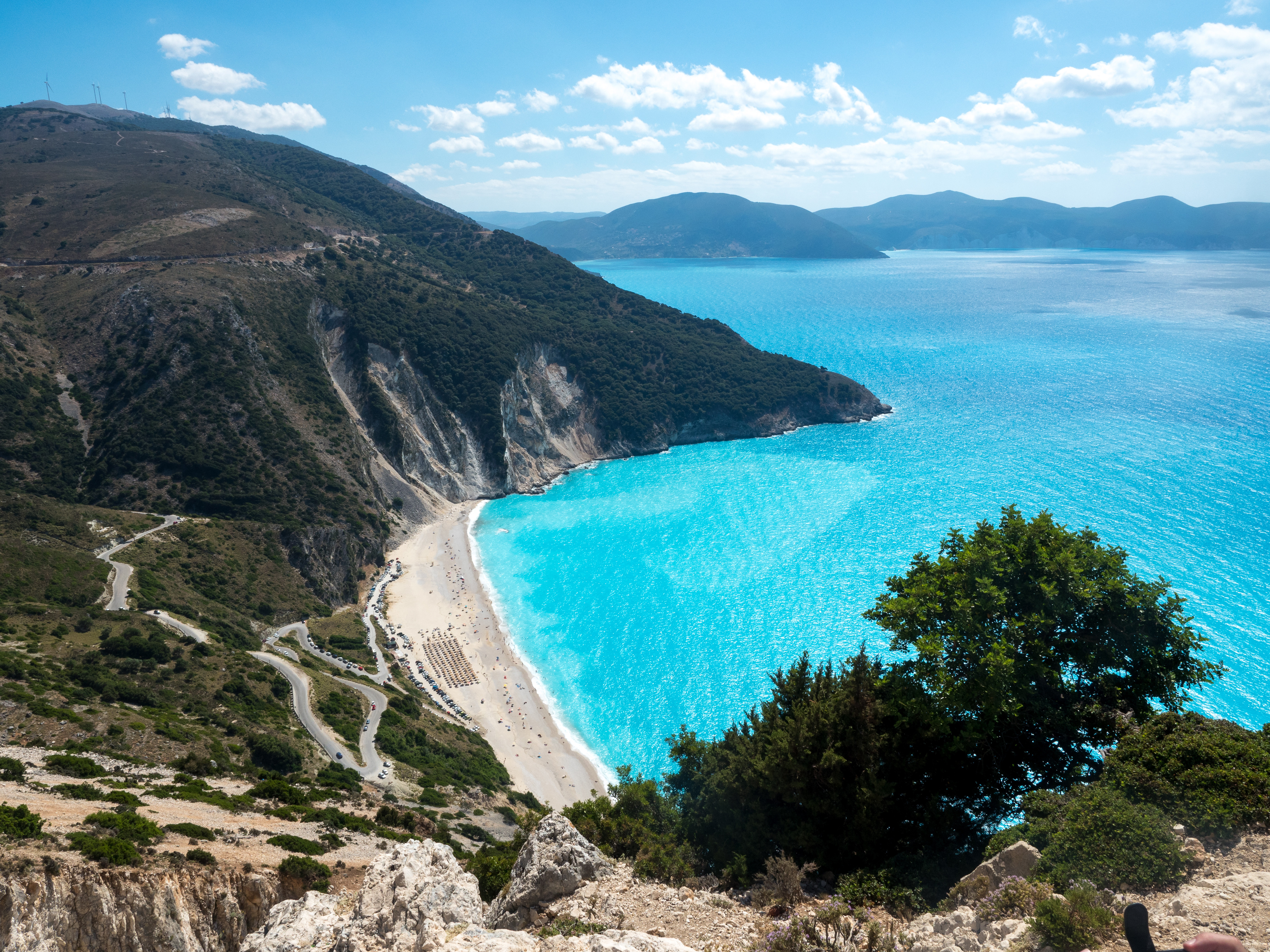 Utsikt over Myrtos-stranden på Kefalonia i Hellas med krystallklart turkis hav, frodige grønne åser og en svingete vei langs kysten på en solfylt dag
