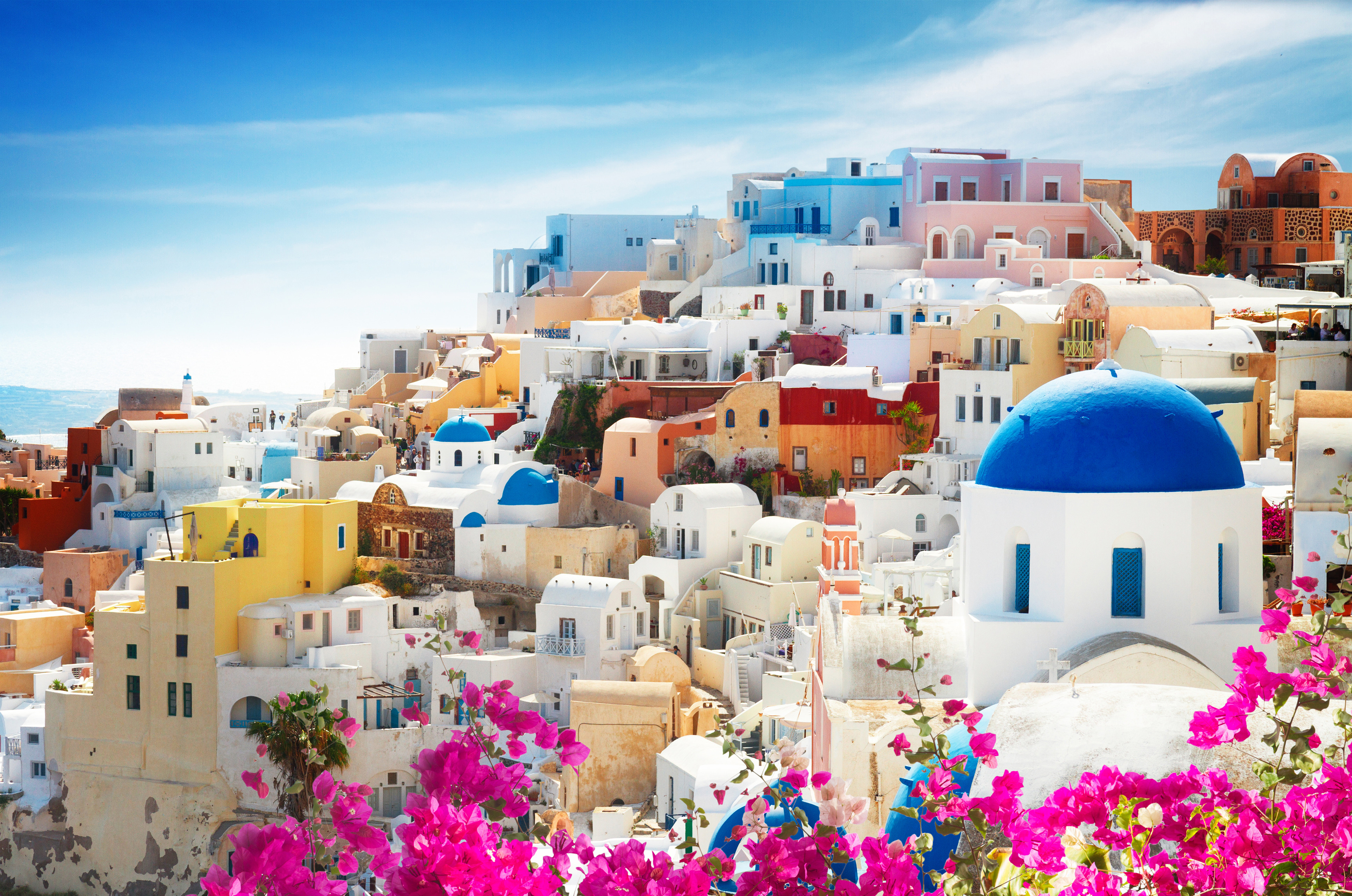 Hvite bygninger med blå tak i Santorini, Hellas, under en klar blå himmel, omgitt av fargerike blomster